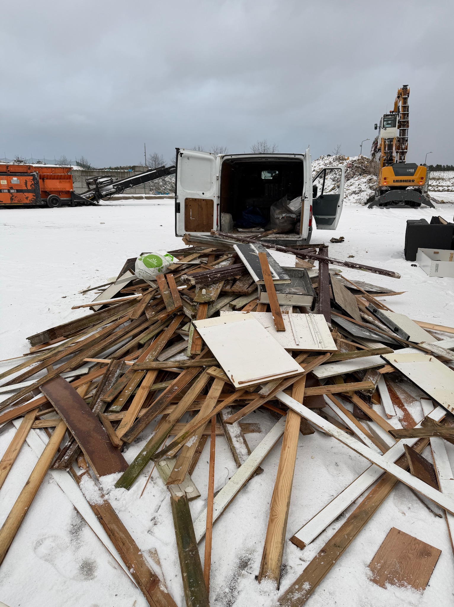 Otwarty furgon na śniegu, za nim sterta gruzu i maszyny budowlane. Widoczne deski, fragmenty mebli i kontener. Pochmurne niebo w tle.