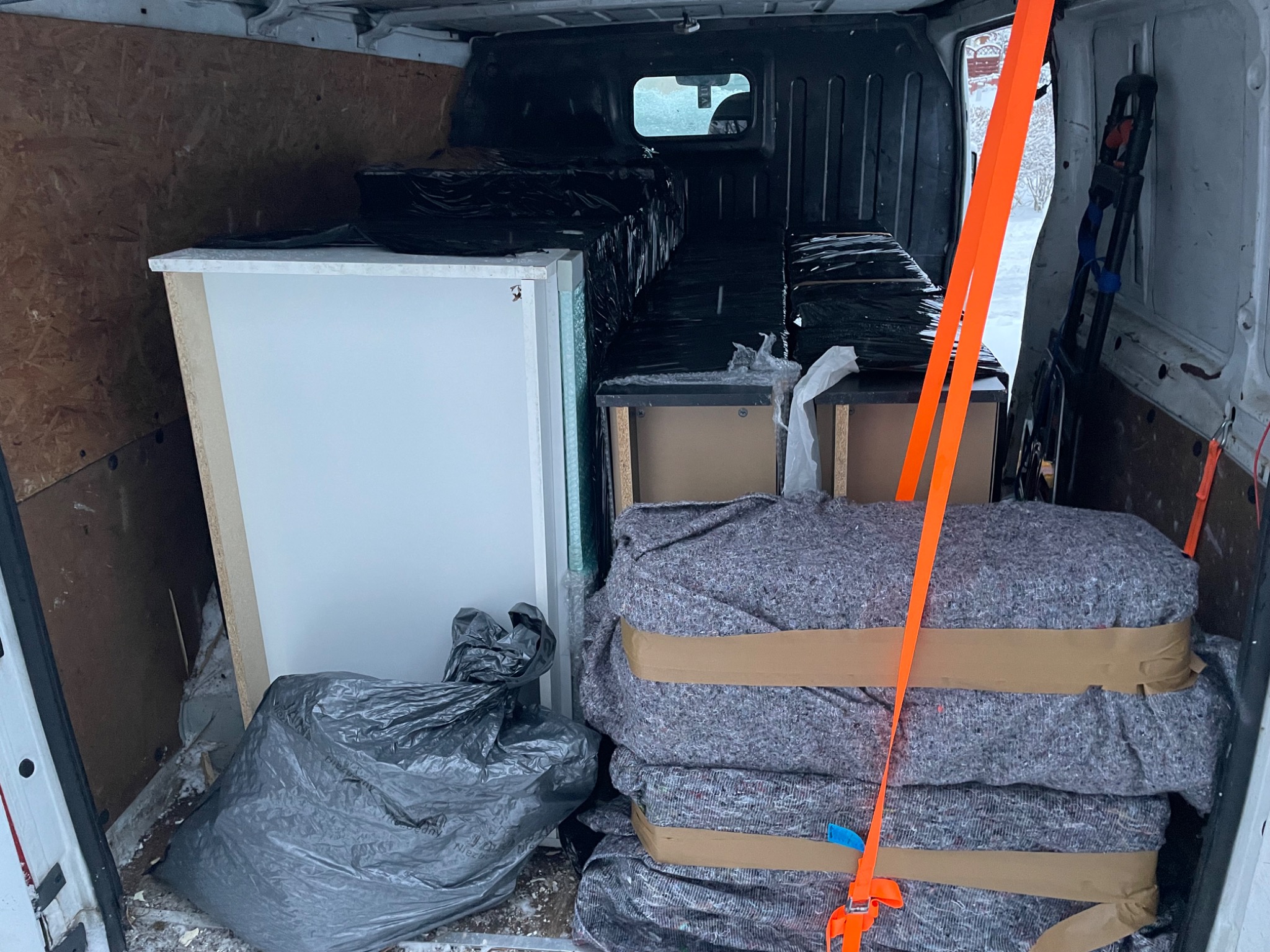 Wnętrze busa załadowane meblami, zabezpieczonymi folią i kocami, gotowe do transportu. Widoczne pasy mocujące i elementy wyposażenia wnętrza pojazdu.