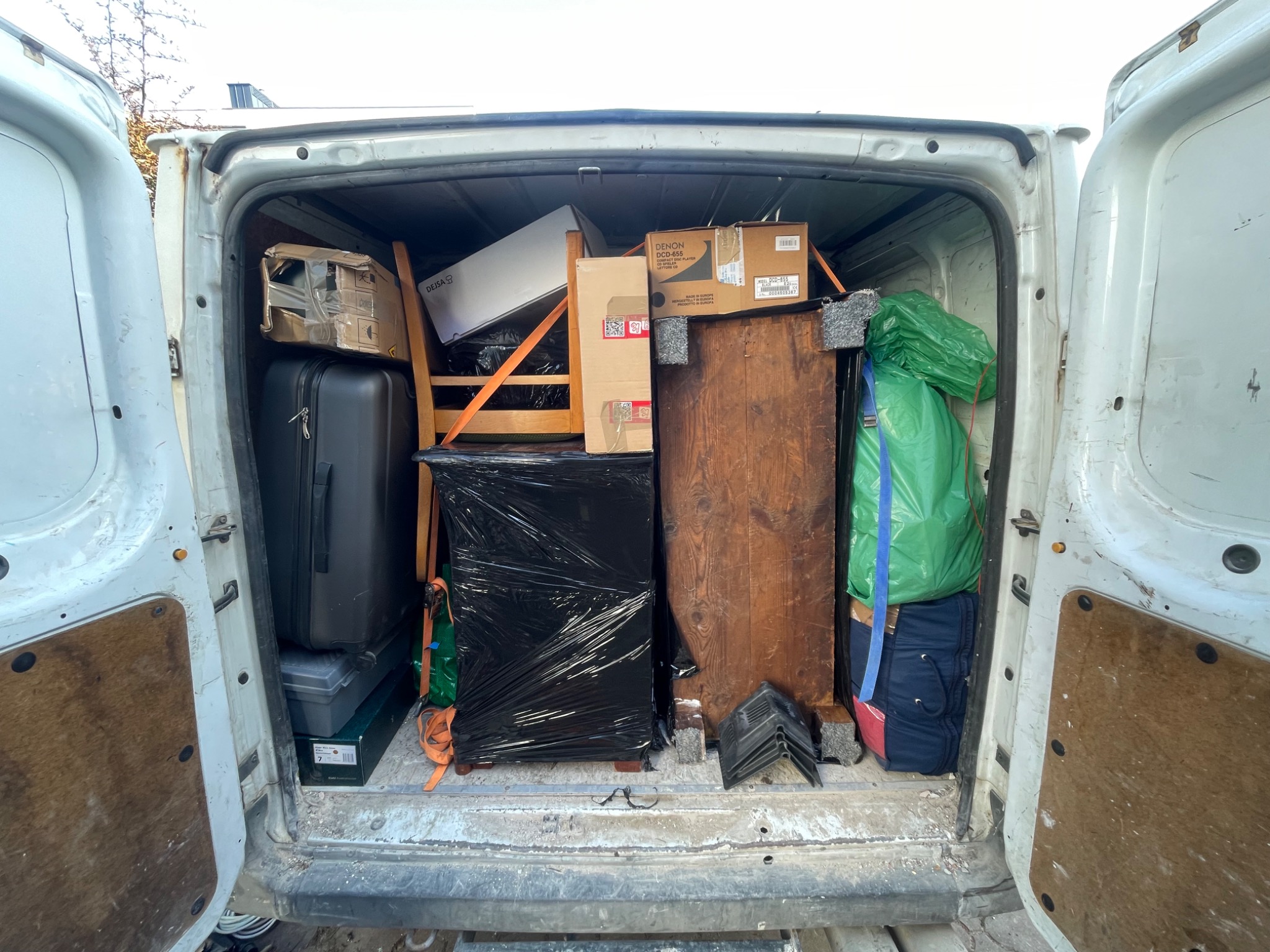 Załadunek busa: meble owinięte folią, kartony i walizka w przestrzeni ładunkowej białego samochodu dostawczego z otwartymi drzwiami.