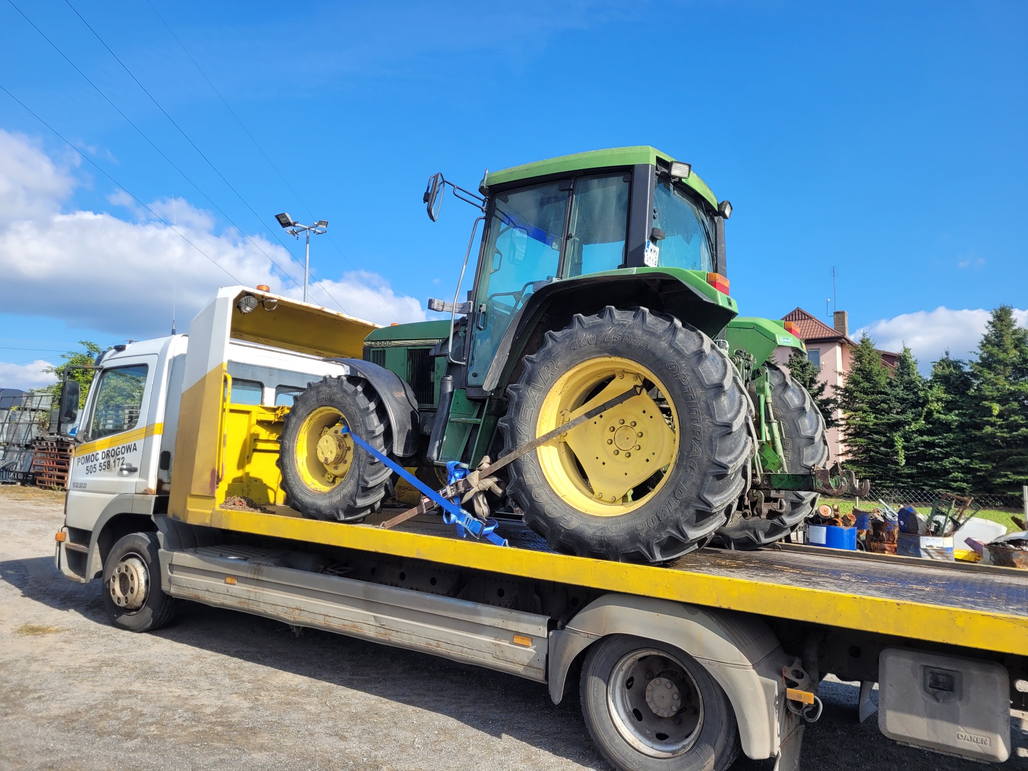 Laweta z zielonym traktorem John Deere na żółtej platformie transportowej, zabezpieczonym niebieskimi pasami. Błękitne niebo w tle.