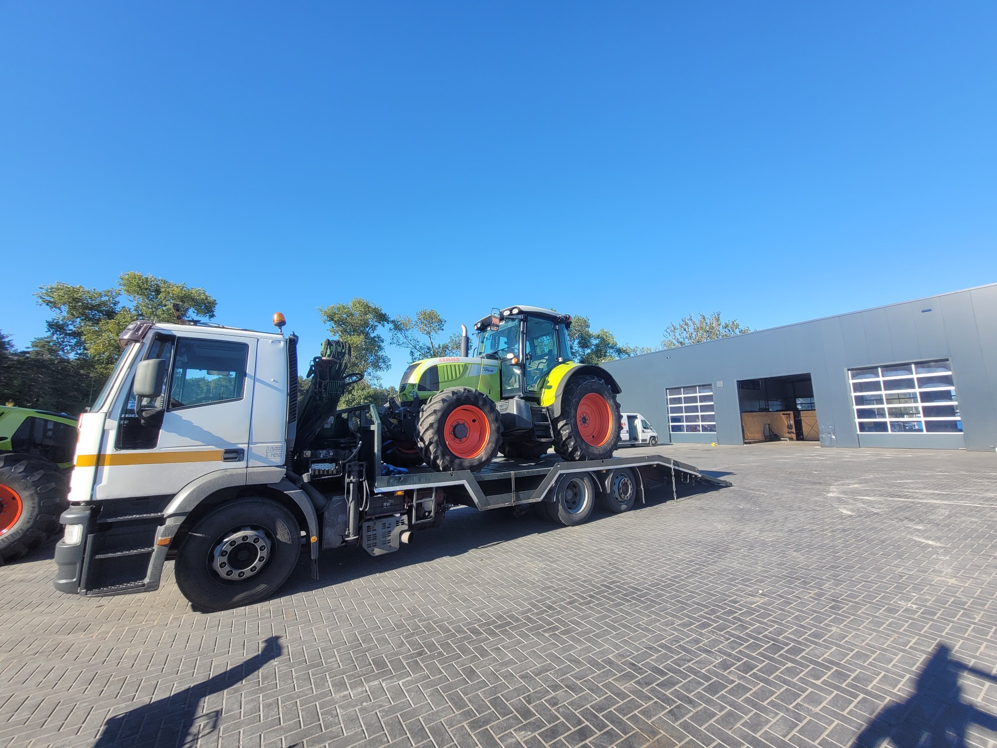 Laweta z traktorem marki Claas Arion na platformie, widok z boku na tle błękitnego nieba i budynku magazynowego z bramami garażowymi.