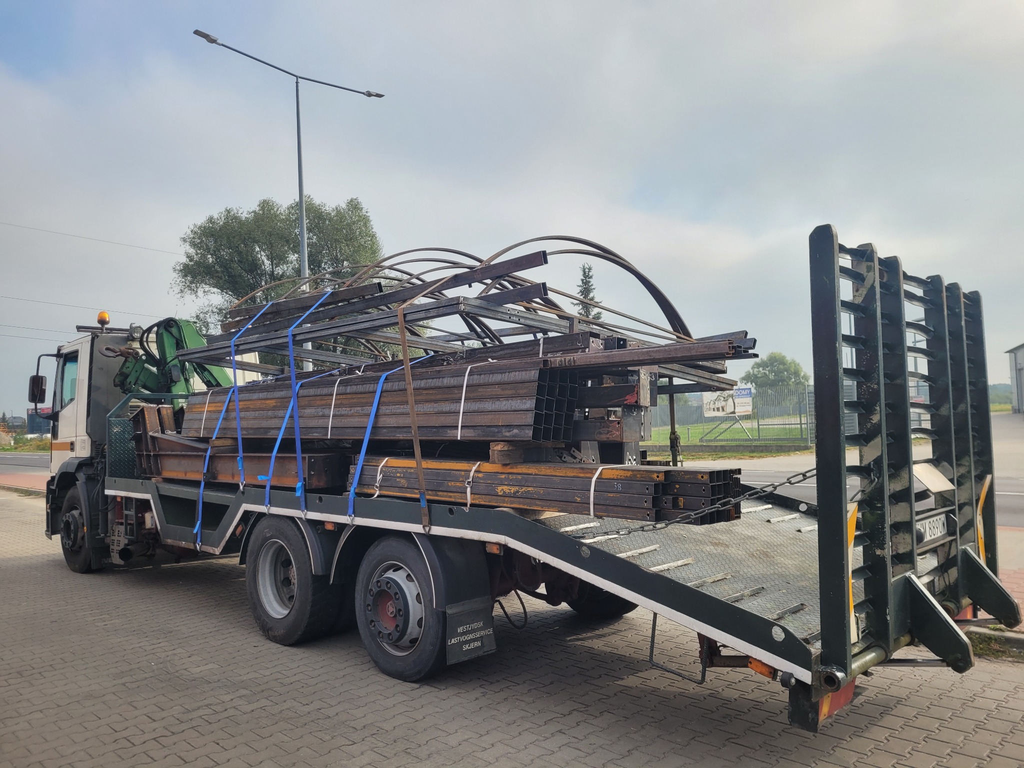 Laweta z ładunkiem metalowych profili i konstrukcji, zabezpieczonych niebieskimi pasami, przygotowana do transportu. Widok z boku, na pierwszym planie kostka brukowa.