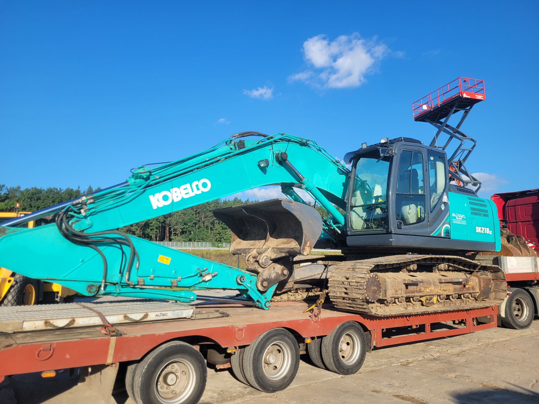 Niebieska koparka Kobelco SK210LC na czerwonej lawecie, widok z boku, błękitne niebo w tle. Sprzęt gotowy do transportu.