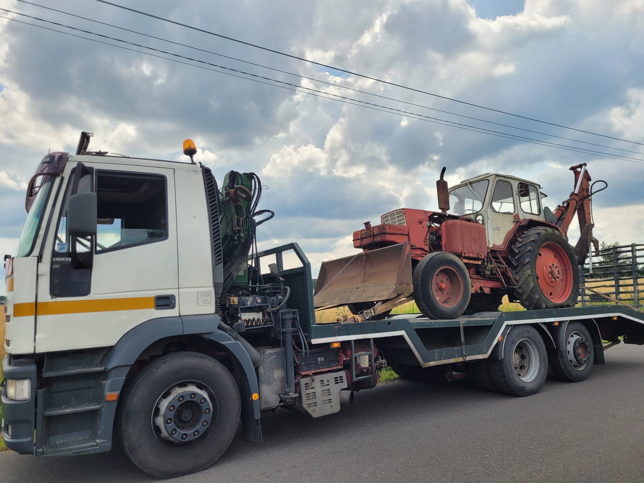 Laweta przewozi stary, czerwono-biały traktor z łyżką na platformie. Widoczny fragment drogi i pola w tle pod pochmurnym niebem.