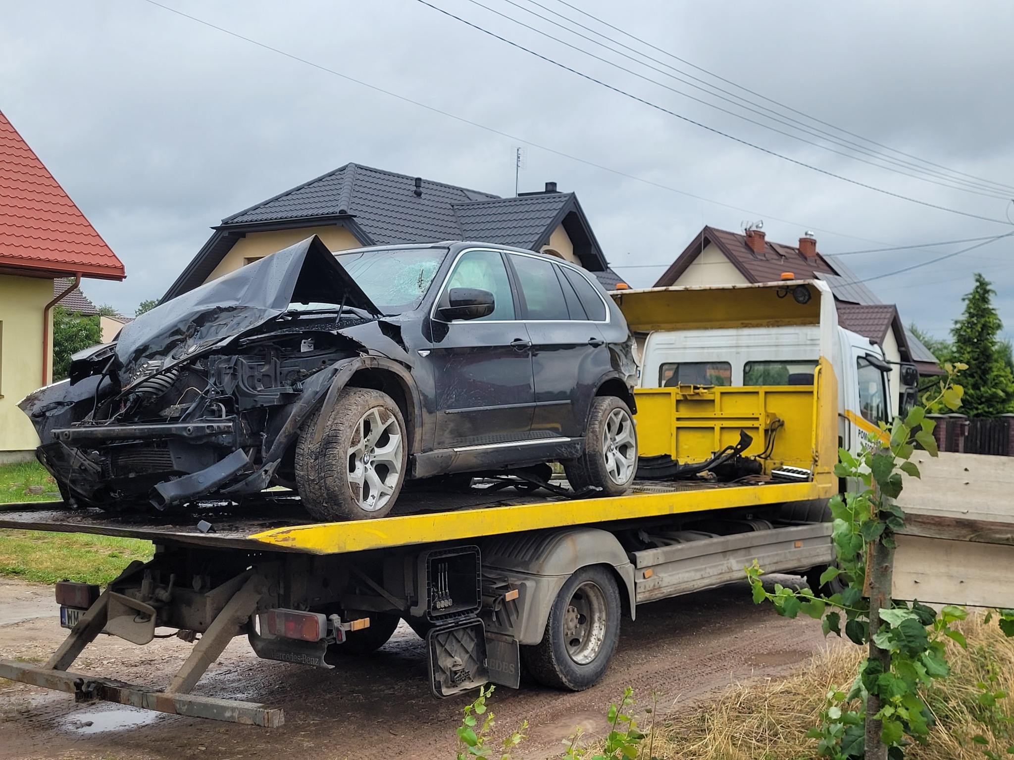 Czarny, uszkodzony SUV na żółtej lawecie, po wypadku, z zniszczonym przodem, na tle budynków. Widok z boku, pochmurne niebo.