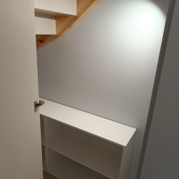 Meblarium Marcin Hałas - Biała szafka z półkami pod schodami, widoczna z otwartymi drzwiami szafy. Drewniane schody i szara ściana w tle. Oświetlenie punktowe podkreśla strukturę ściany.
