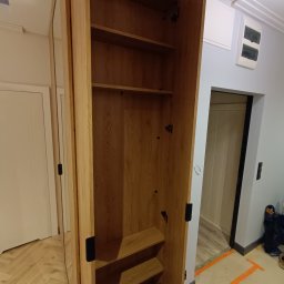 Meblarium Marcin Hałas - Otwarta, drewniana szafa z półkami i lustrzanym frontem, widoczna w jasnym wnętrzu z parkietem ułożonym w jodełkę. Drzwi szafy otwarte, ukazują wnętrze z półkami.