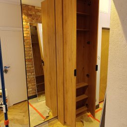 Meblarium Marcin Hałas - Montaż szafy z lustrem w ramie. Otwarta szafa z półkami, widoczne wnętrze. Podłoga zabezpieczona taśmą. Ceglana ściana odbija się w lustrze. Ujęcie z bliska.