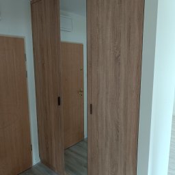 Meblarium Marcin Hałas - Trzydrzwiowa szafa z lustrem w środkowej części, wykończona okleiną imitującą drewno. Widoczne dwie drewniane drzwi po bokach i fragment podłogi.