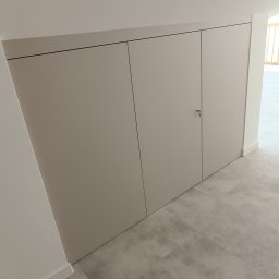 Meblarium Marcin Hałas - Jasna szafa wnękowa z trzema drzwiami, zamykana na klucz, wkomponowana w skos dachu, na tle szarej podłogi i białych ścian.