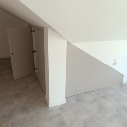 Meblarium Marcin Hałas - Biała szafa wnękowa z otwartymi drzwiami, dopasowana do skosu dachu, na szarej podłodze. Widoczne zawiasy i wnętrze szafy.