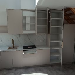 Meblarium Marcin Hałas - Jasna kuchnia na wymiar z marmurowym blatem i złotą armaturą. Częściowo otwarte szafki ukazują puste półki. Na podłodze widoczny cień.