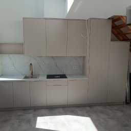 Meblarium Marcin Hałas - Jasna kuchnia na wymiar z marmurowym blatem i złotą armaturą, widok na drewniane schody i okno dachowe, minimalistyczny design.