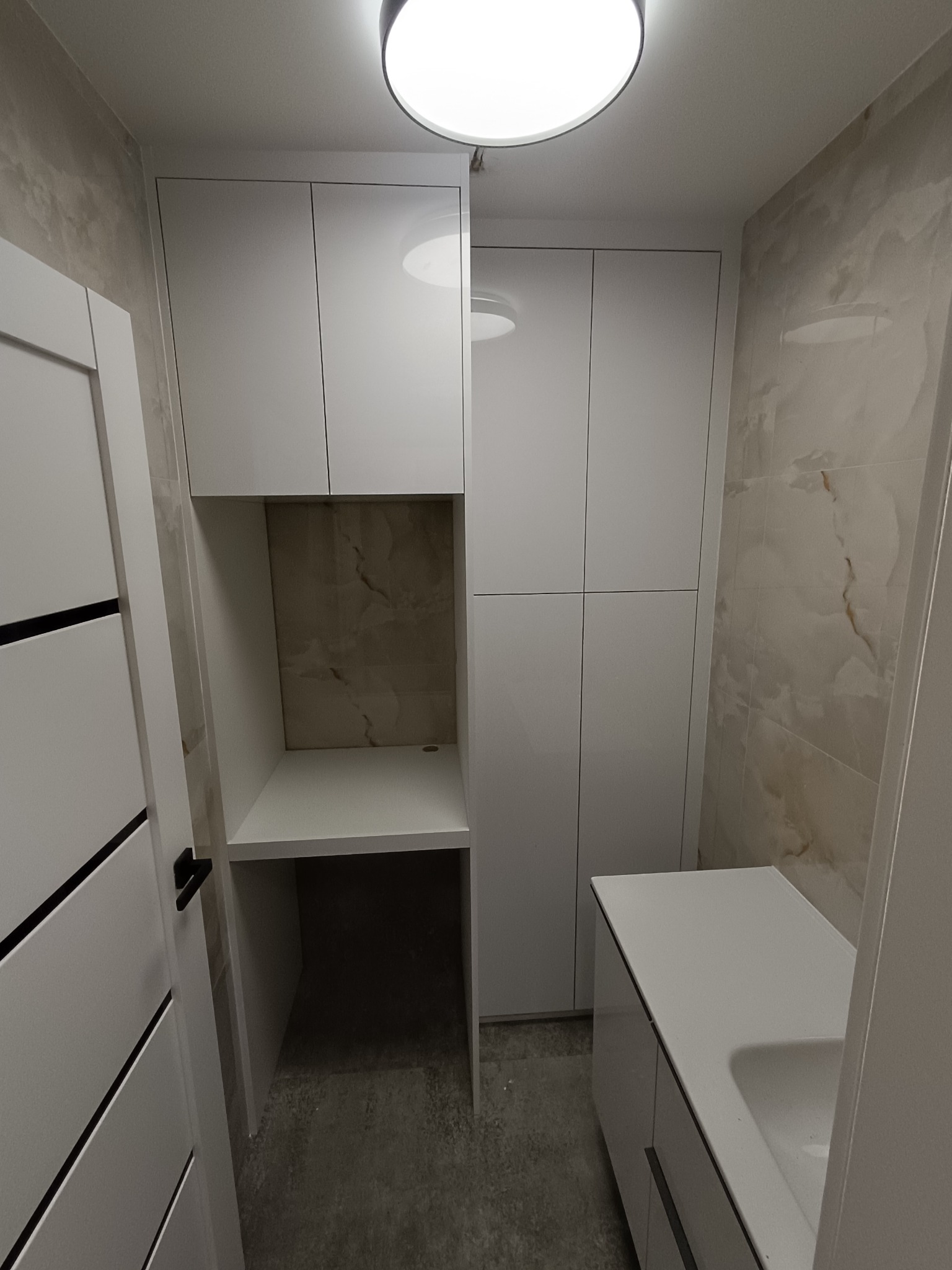 Biała zabudowa meblowa w łazience z szafkami wiszącymi, blatem roboczym i szafką pod umywalką. Minimalistyczny design, jasne wnętrze, płytki imitujące marmur na ścianach.