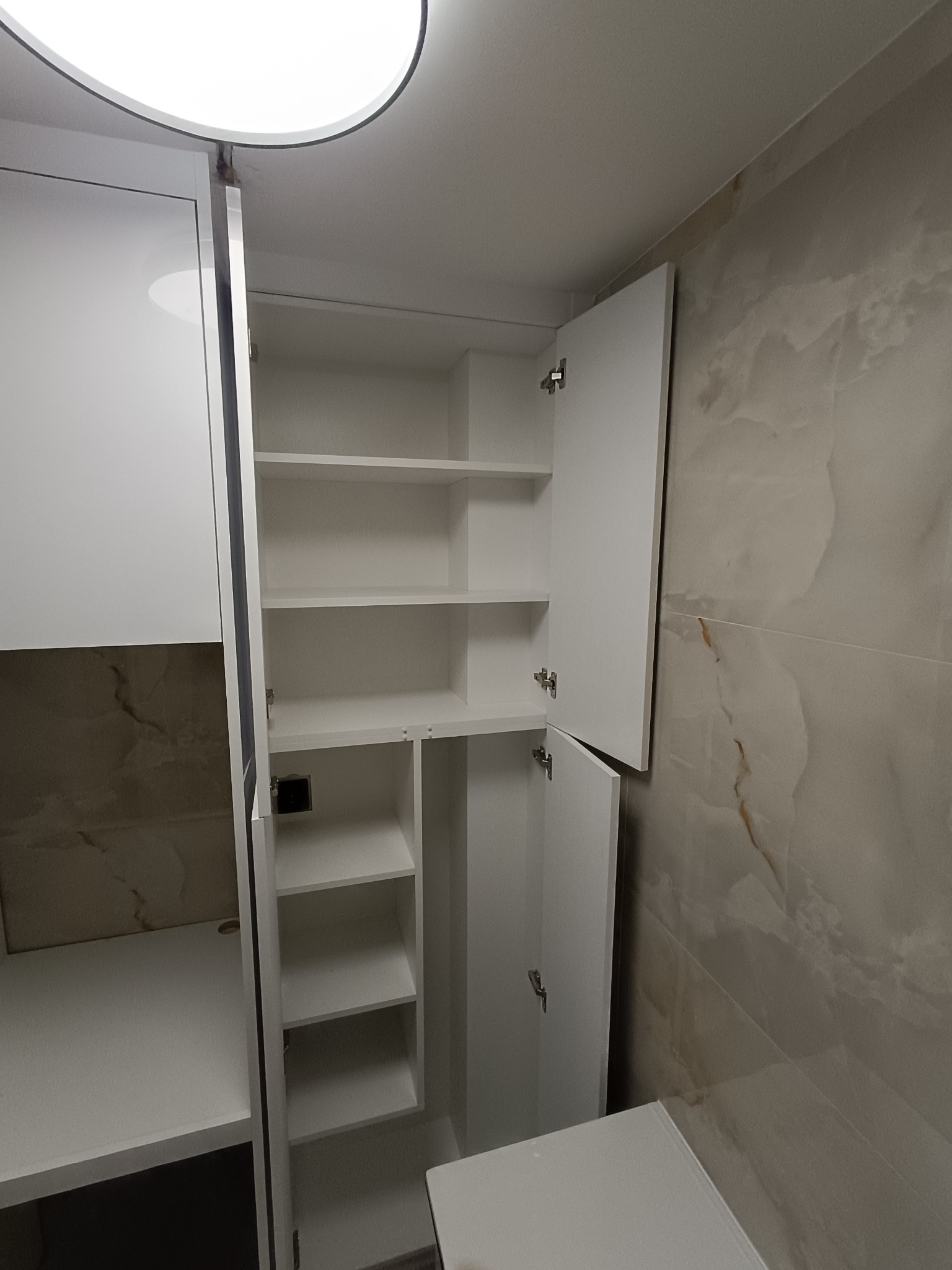 Biała szafa wnękowa z otwartymi drzwiami, widoczne półki. Minimalistyczny design, jasne wnętrze, marmurowe płytki w tle. Oświetlenie sufitowe.