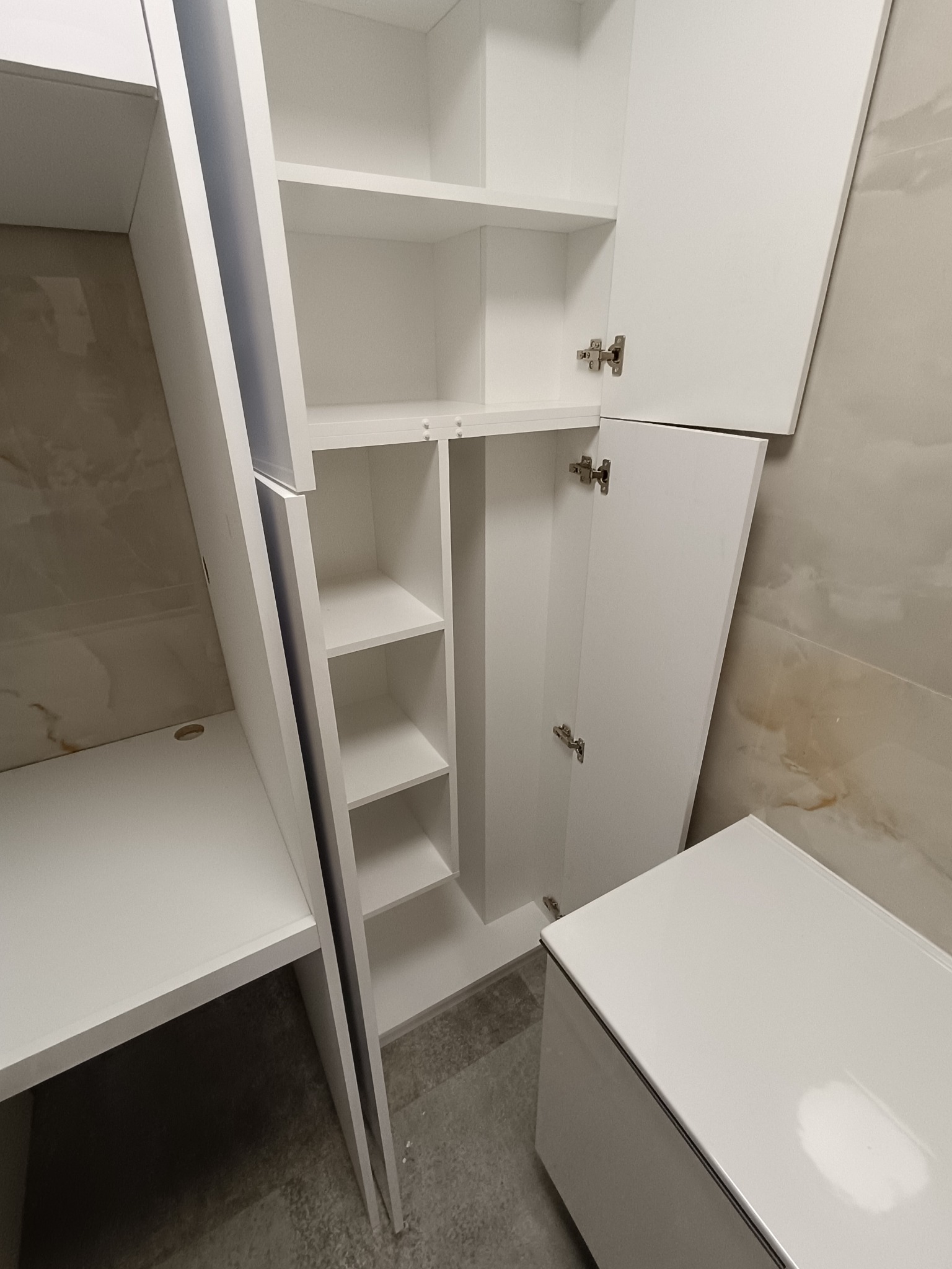 Biała szafa wnękowa z otwartymi drzwiami i półkami, zintegrowana z biurkiem, na tle beżowej ściany. Minimalistyczny design, nowoczesne wykończenie.