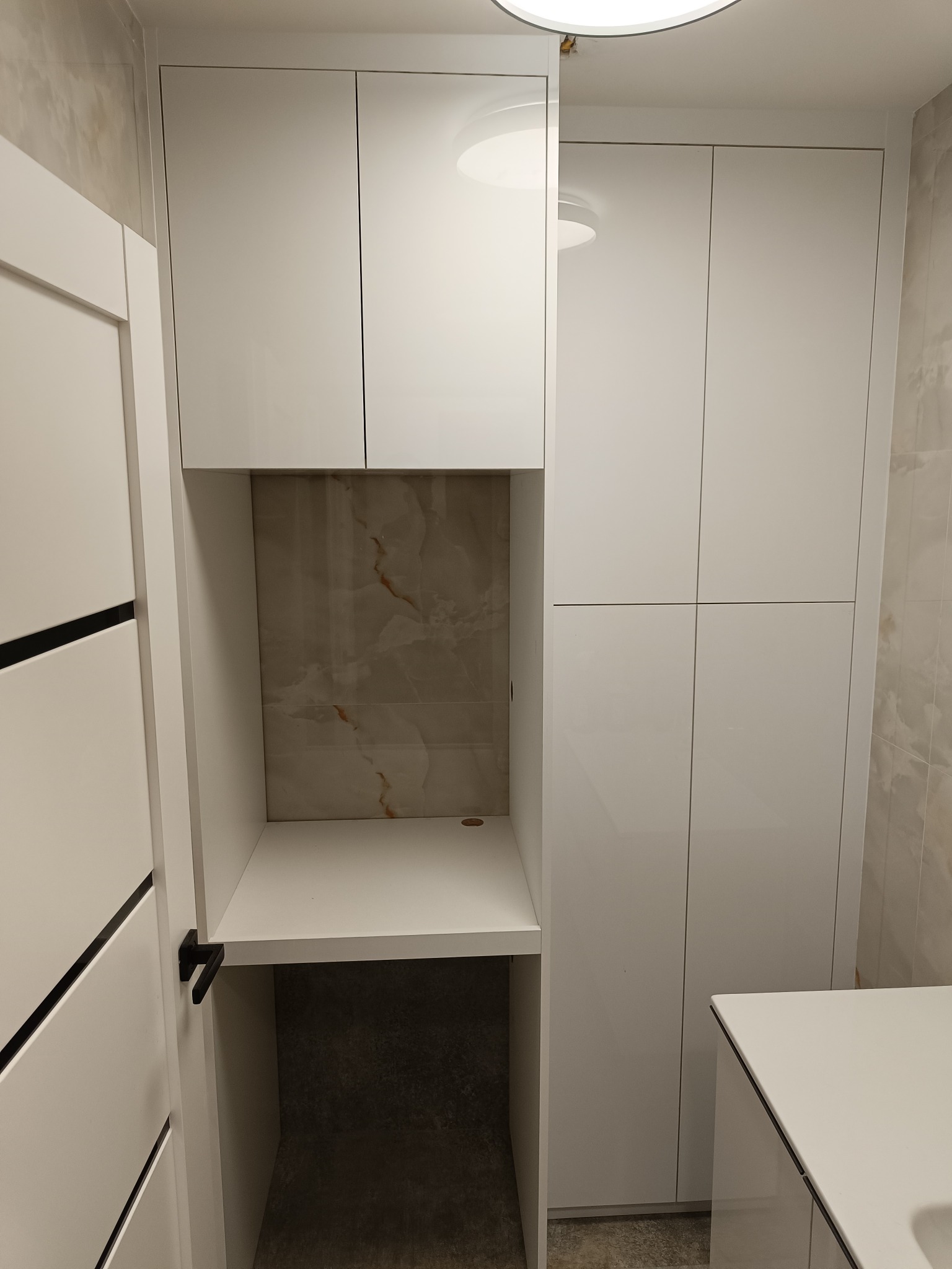 Biała zabudowa meblowa na wymiar w łazience z blatem i szafkami. Minimalistyczny design, jasne wnętrze, marmurowe wykończenie ściany nad blatem. Nowoczesne rozwiązanie.