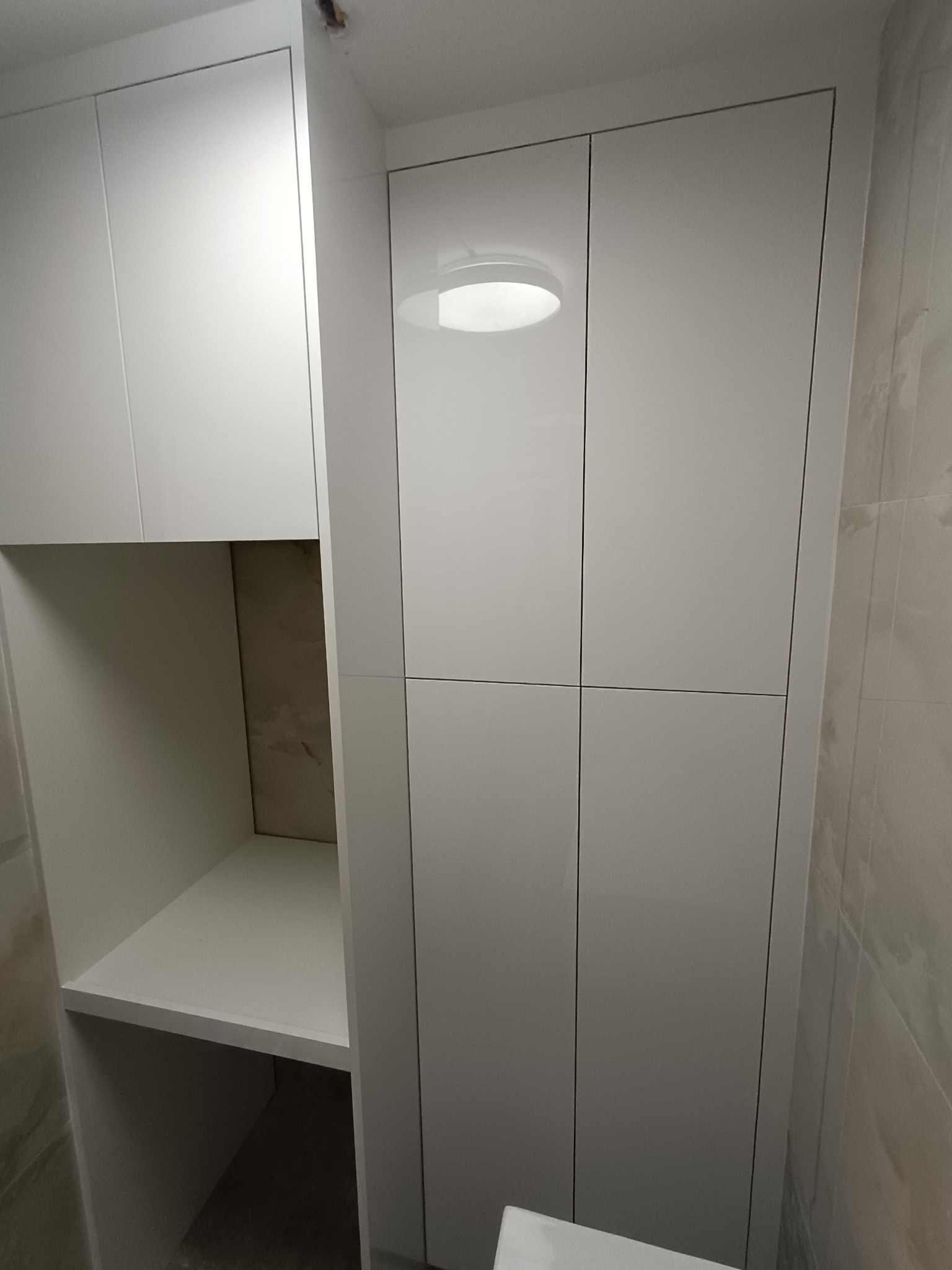 Biała szafa wnękowa z połyskiem, z otwartą półką po lewej stronie. Minimalistyczny design, dopasowana do przestrzeni łazienki z jasnymi płytkami na ścianie.