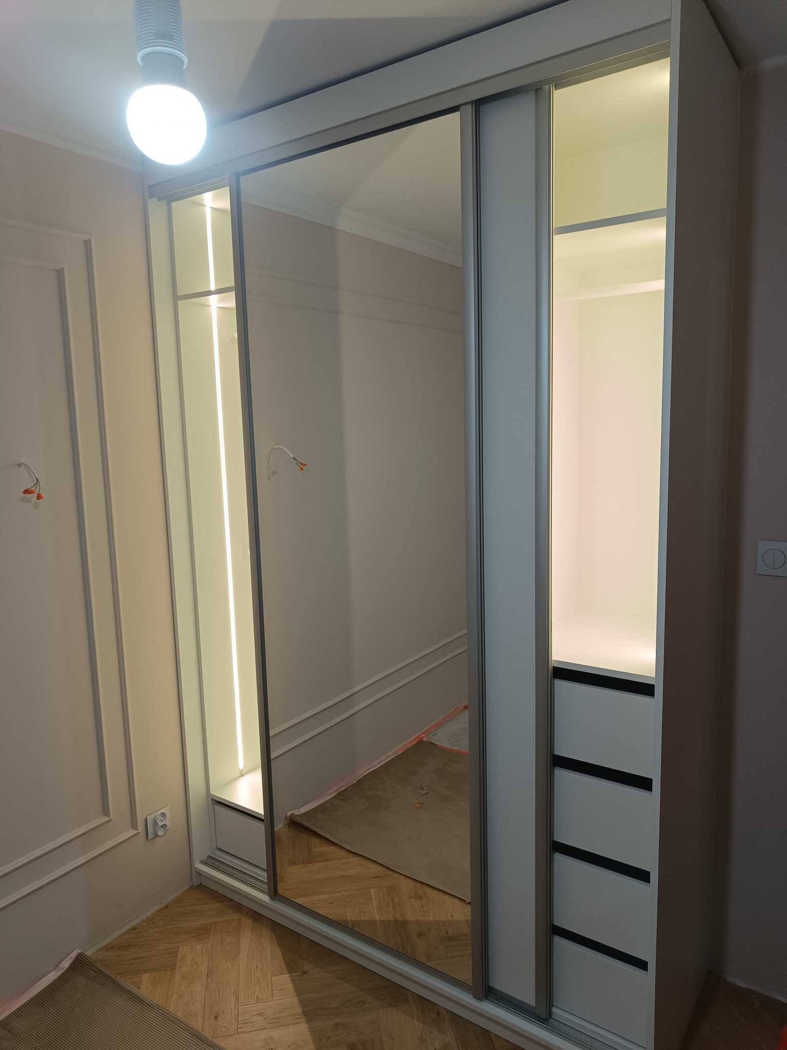 Biała szafa przesuwna z lustrem i oświetleniem LED, wbudowana w ścianę z dekoracyjnymi listwami. Widoczne półki i szuflady wewnątrz szafy.