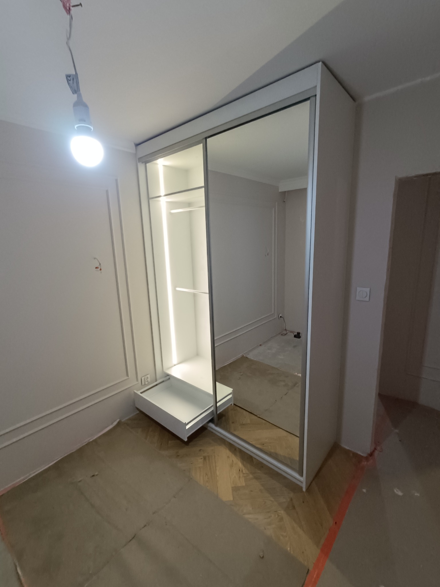 Biała szafa wnękowa z przesuwnymi drzwiami lustrzanymi i oświetleniem LED wewnątrz, widoczna w trakcie montażu, z zabezpieczoną podłogą.