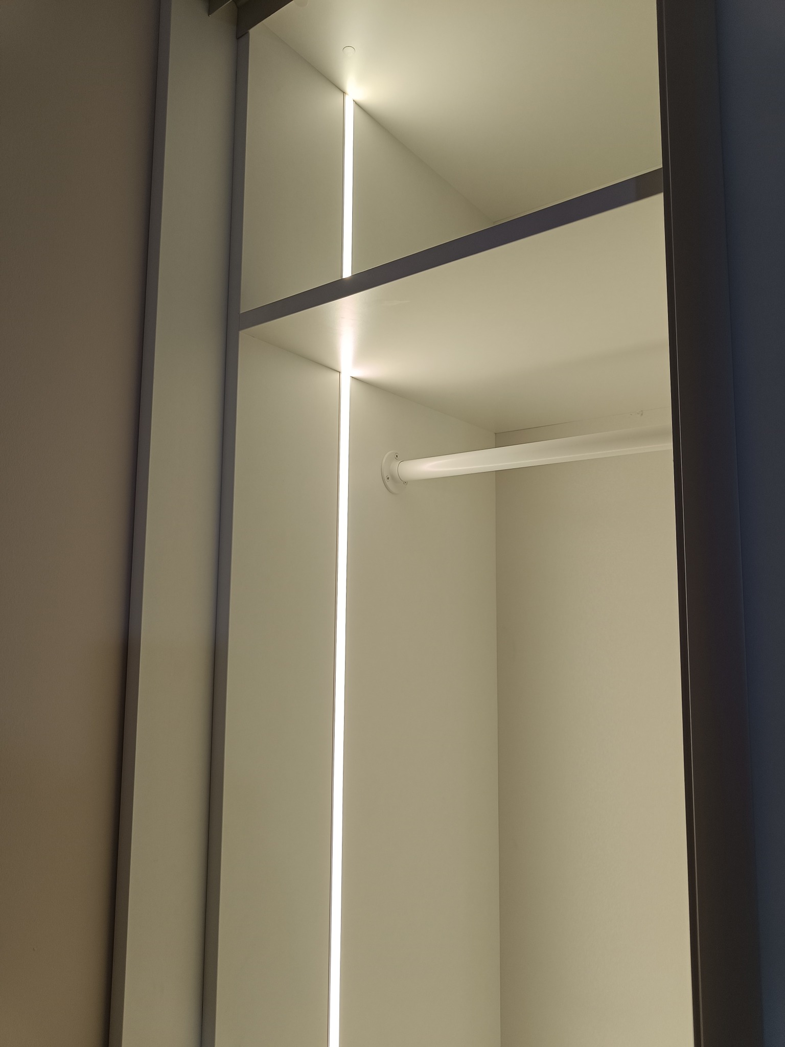Wnętrze szafy wnękowej z oświetleniem LED, widoczny drążek na ubrania i półka. Minimalistyczny design, jasne kolory, nowoczesne rozwiązanie oświetleniowe.