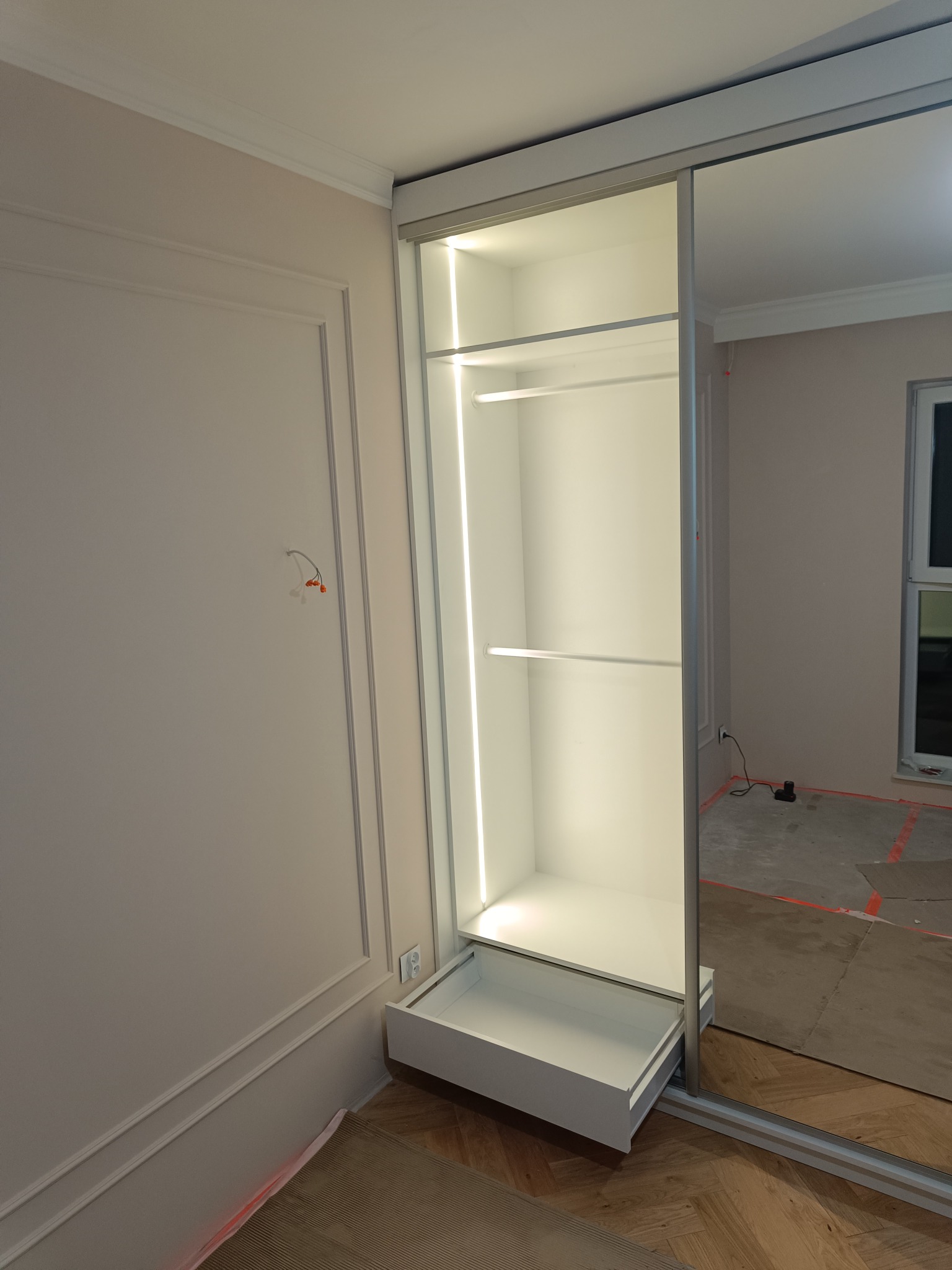 Wnętrze szafy wnękowej z oświetleniem LED i wysuwaną szufladą, widoczne odbicie w lustrzanych drzwiach przesuwnych. Ściana z dekoracyjną sztukaterią.