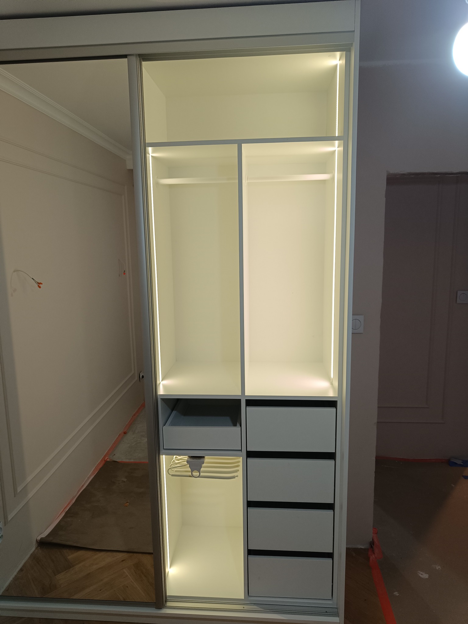 Biała szafa wnękowa z przesuwnymi drzwiami i oświetleniem LED, widoczne półki, szuflady i wieszak na spodnie. Minimalistyczny design, wnętrze szafy.