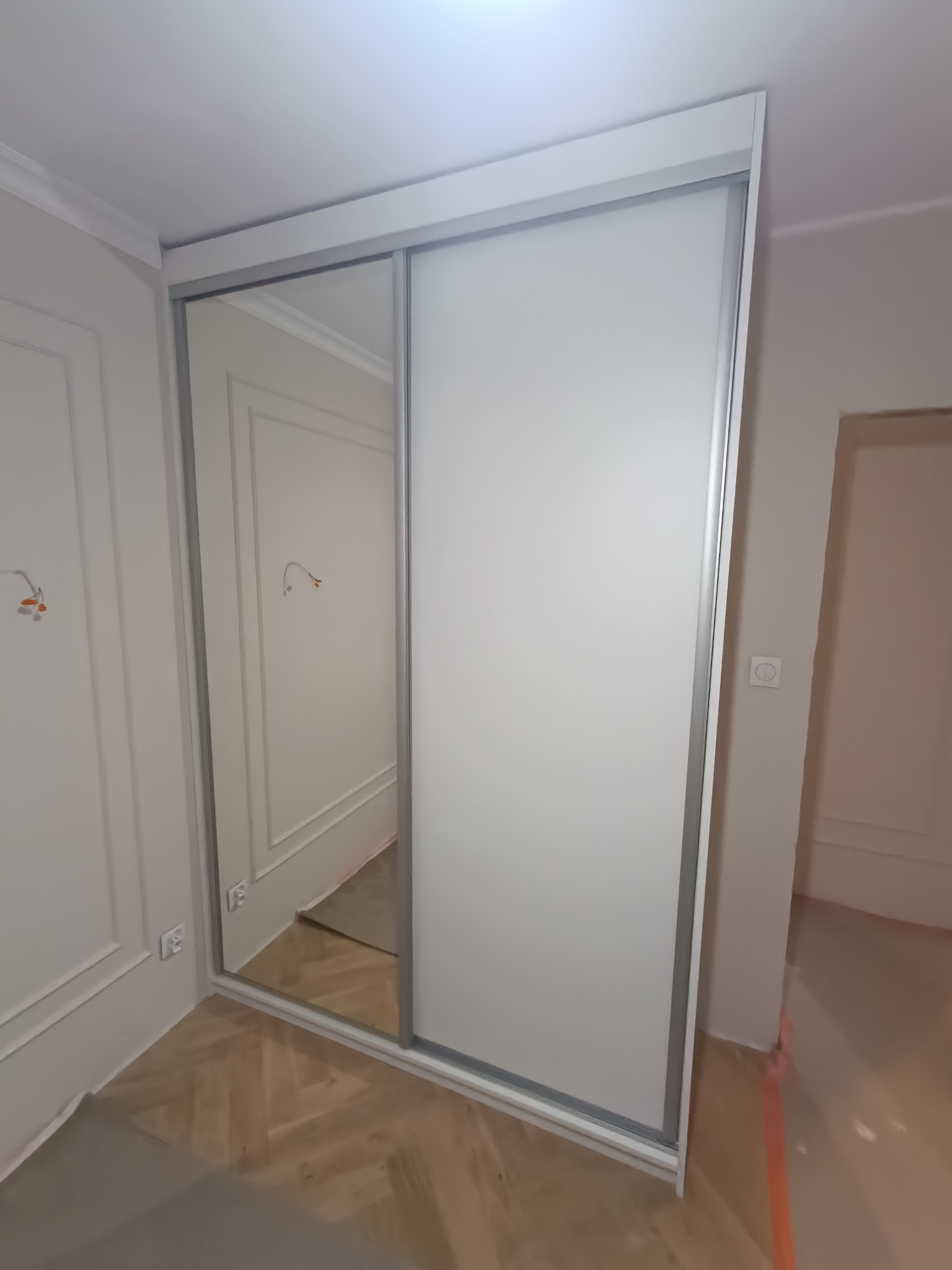 Biała szafa przesuwna z lustrem wnękowym, widoczna na tle ściany z dekoracyjnymi listwami i parkietem. Minimalistyczny design, srebrne obramowanie drzwi.