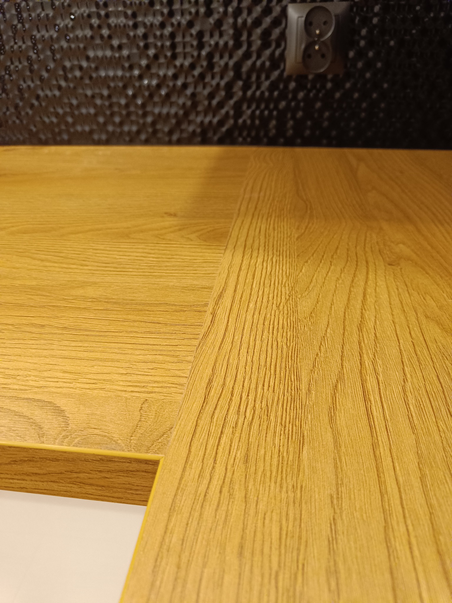 Fragment blatu kuchennego z wyraźnym rysunkiem drewna, na tle czarnej ściany z gniazdkiem elektrycznym. Ujęcie z bliska, ukazujące detale wykończenia.