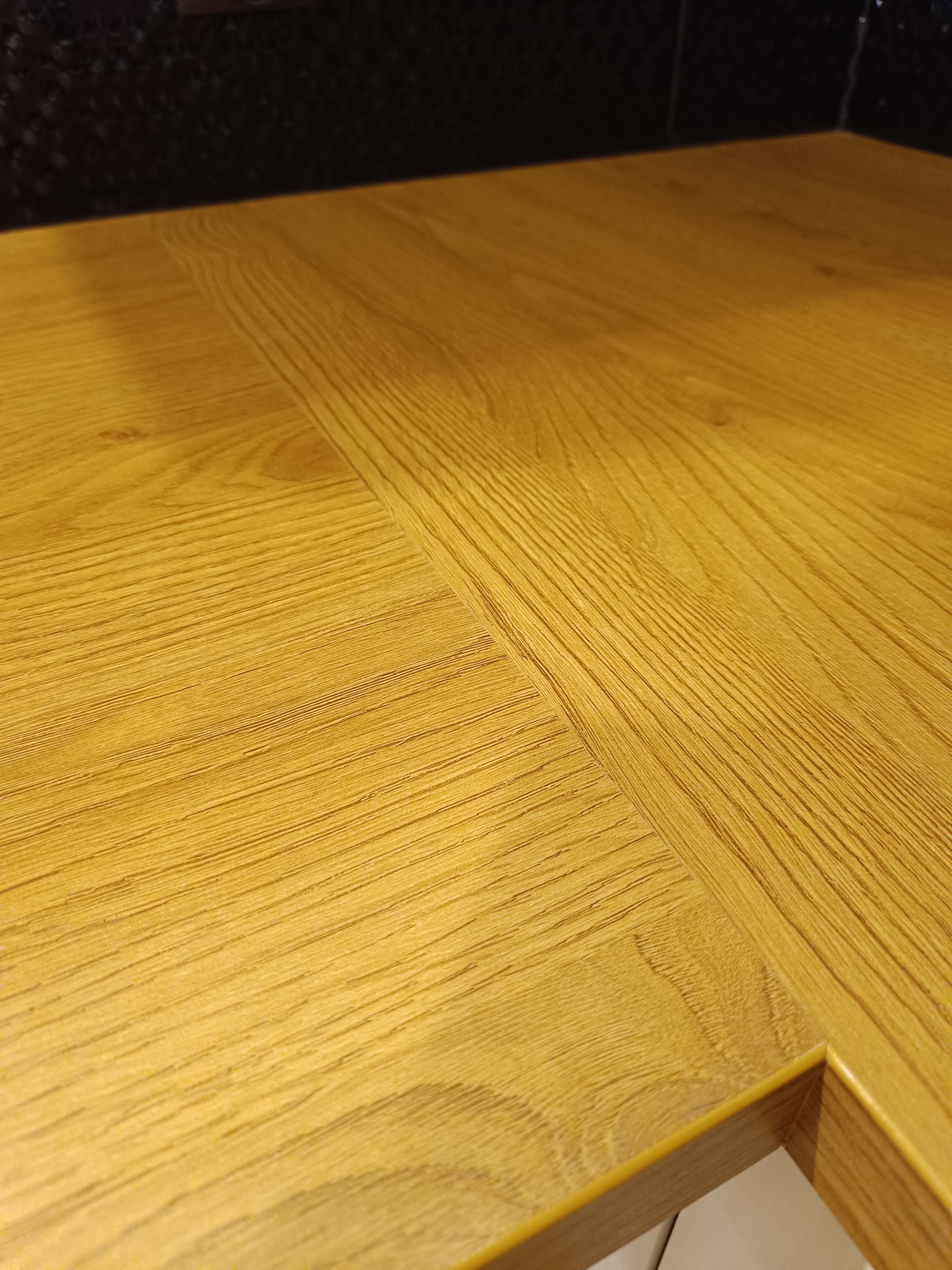 Blat kuchenny z jasnego drewna z widocznym łączeniem i wycięciem, na tle ciemnej ściany. Ujęcie z bliska, ukazujące strukturę drewna i detale wykończenia.