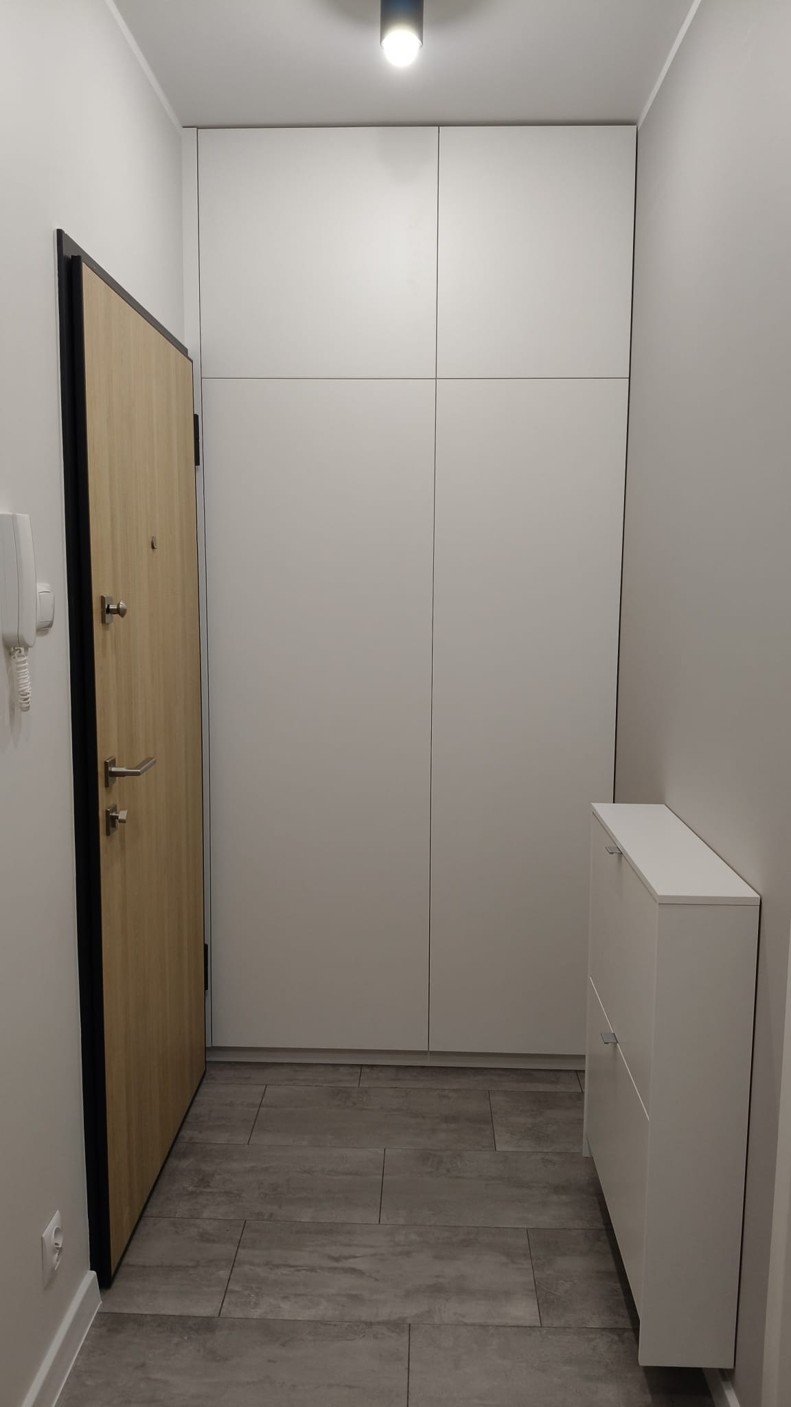 Jasna szafa wnękowa z frontami bez uchwytów, dopasowana do wysokości pomieszczenia, obok drzwi wejściowych i szafki na buty w przedpokoju z szarą podłogą.
