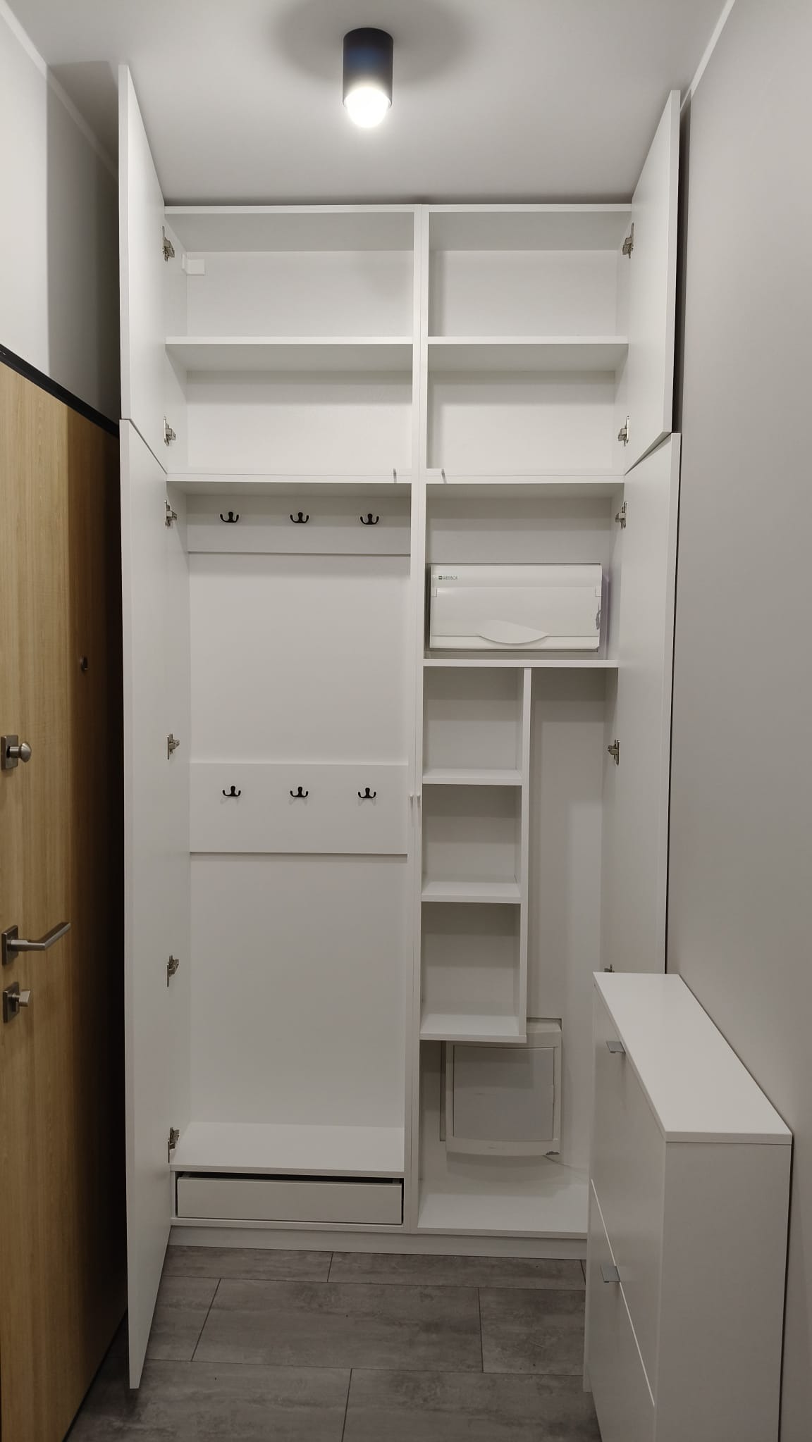 Biała szafa wnękowa z otwartymi drzwiami, widoczne półki, wieszaki i szuflada. Minimalistyczny design, szare płytki podłogowe, oświetlenie sufitowe.