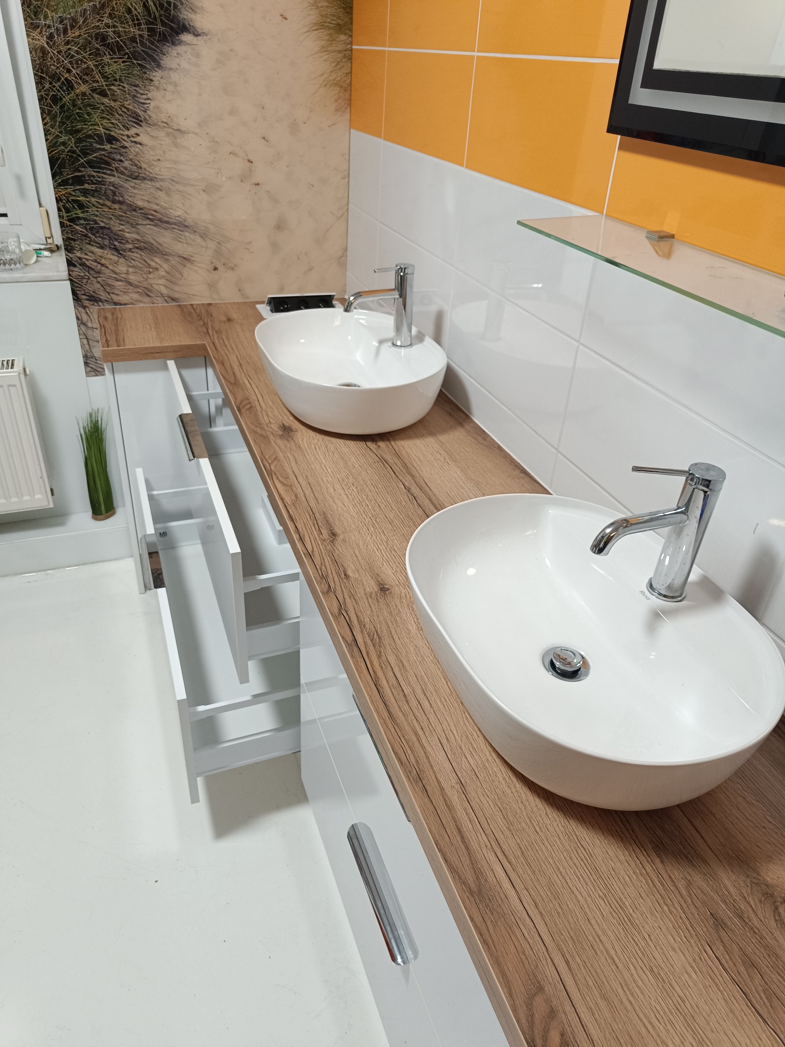 Nowoczesna łazienka z dwoma owalnymi umywalkami nablatowymi na drewnianym blacie. Białe szafki z otwartymi szufladami, żółte płytki i dekoracja z motywem plaży.