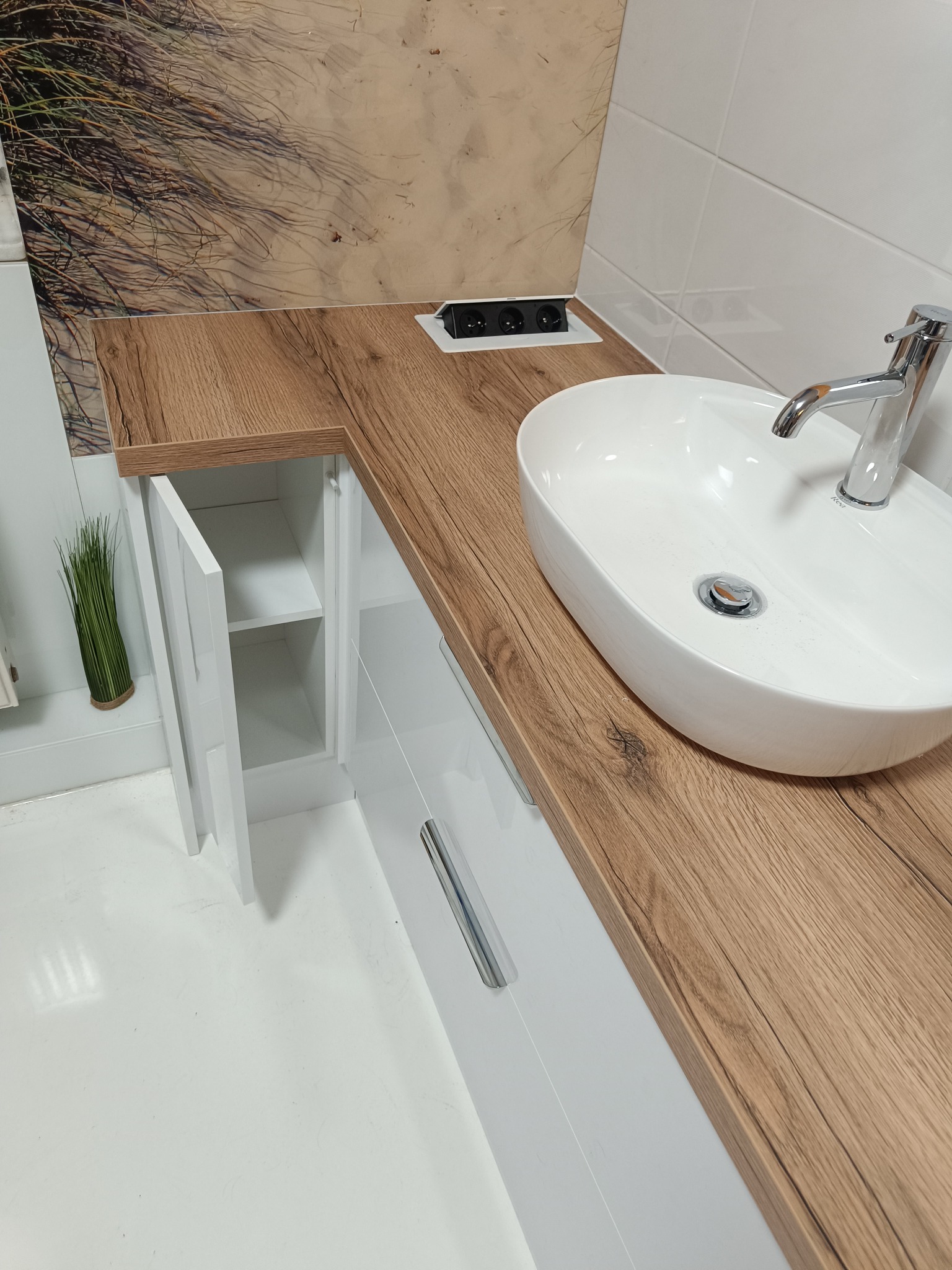 Nowoczesna szafka łazienkowa z drewnianym blatem, owalną umywalką i otwartą szafką boczną. Nad blatem widoczny panel z gniazdkami elektrycznymi.