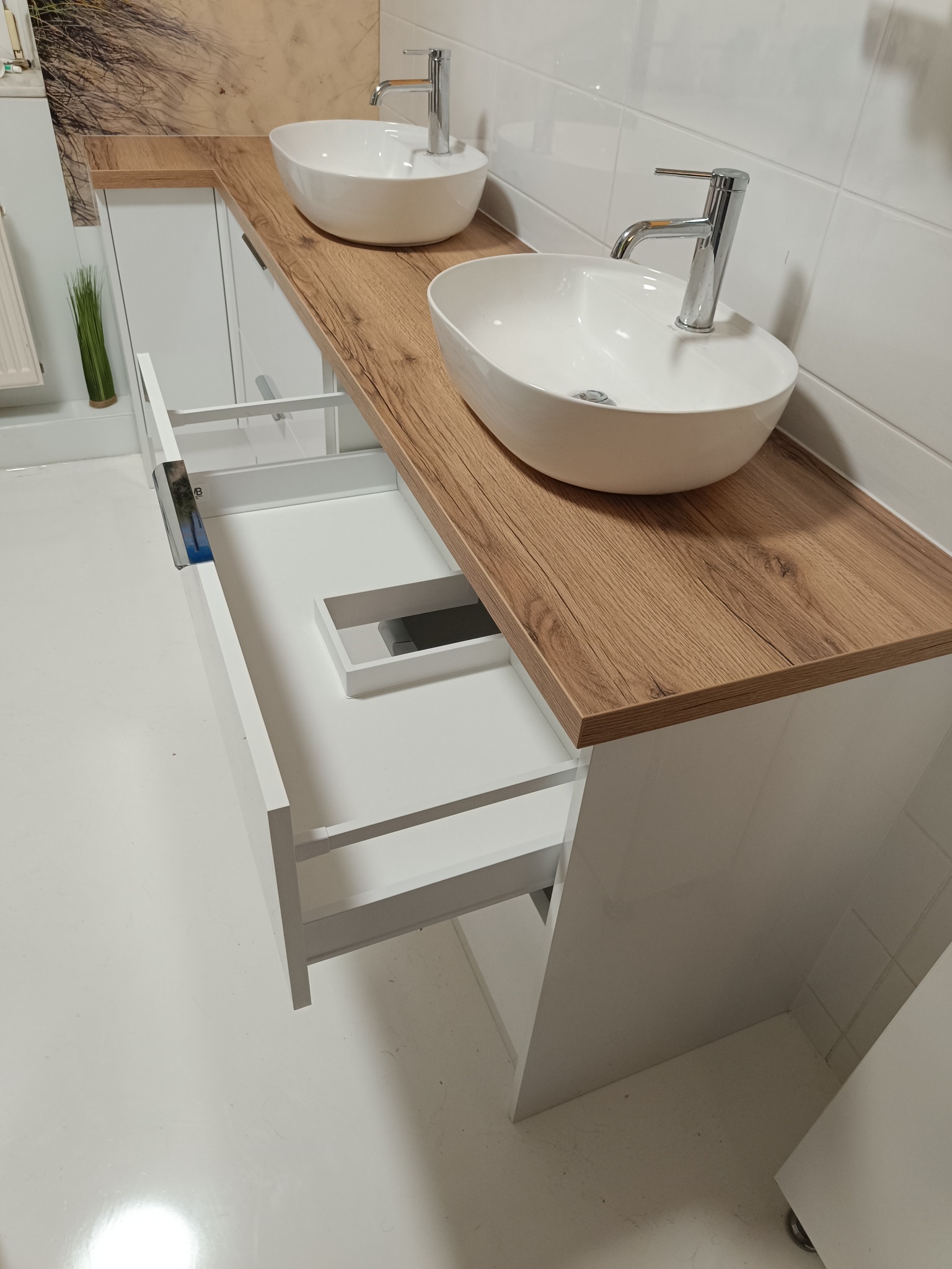 Nowoczesna łazienka z białymi szafkami na wymiar, drewnianym blatem i owalnymi umywalkami. Otwarta szuflada ukazuje wnętrze szafki. Minimalistyczny design.