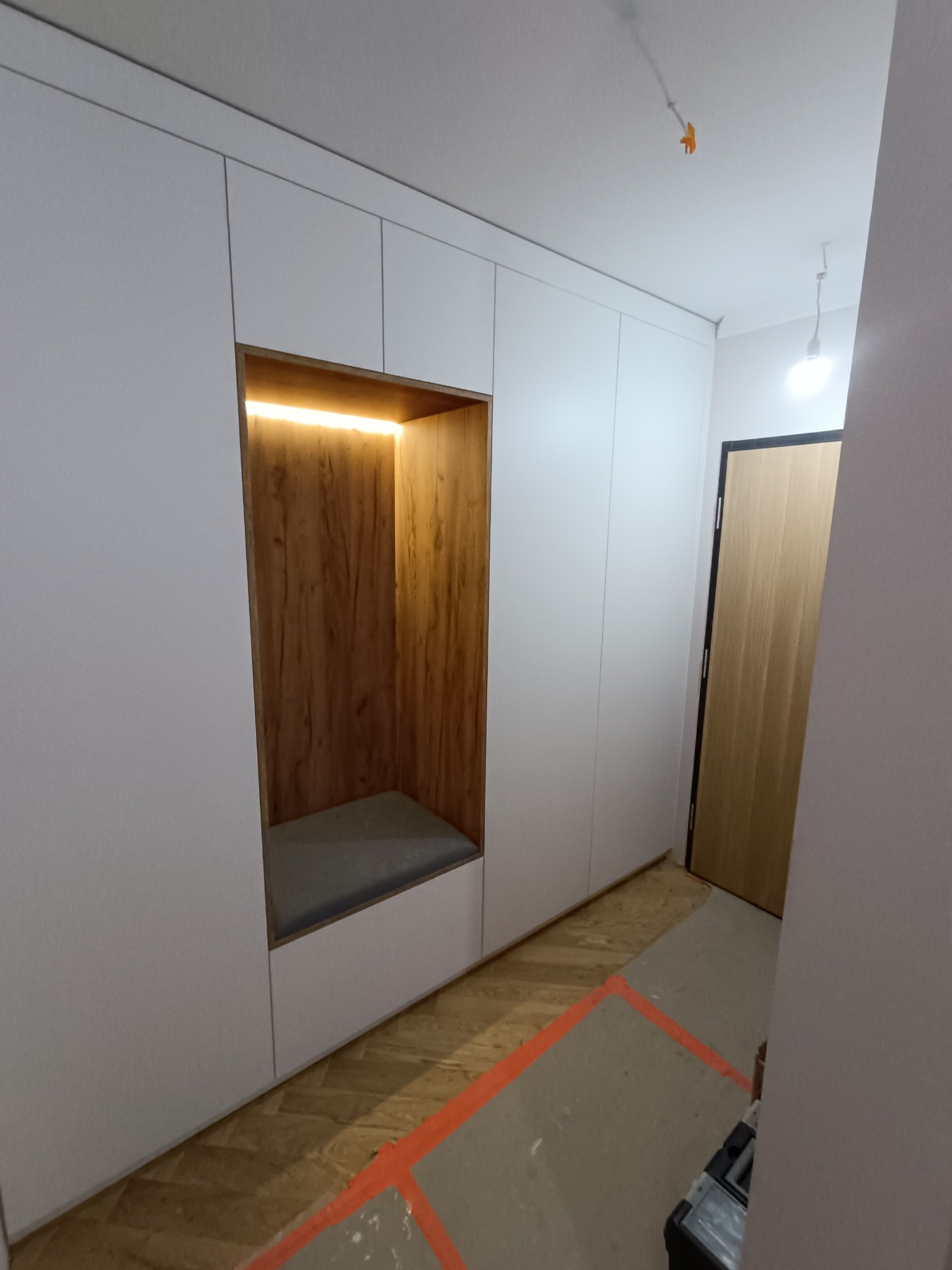 Biała szafa wnękowa z drewnianą wnęką oświetloną LED, z siedziskiem. Widoczny fragment podłogi i drzwi. Minimalistyczny design, nowoczesne wykończenie.