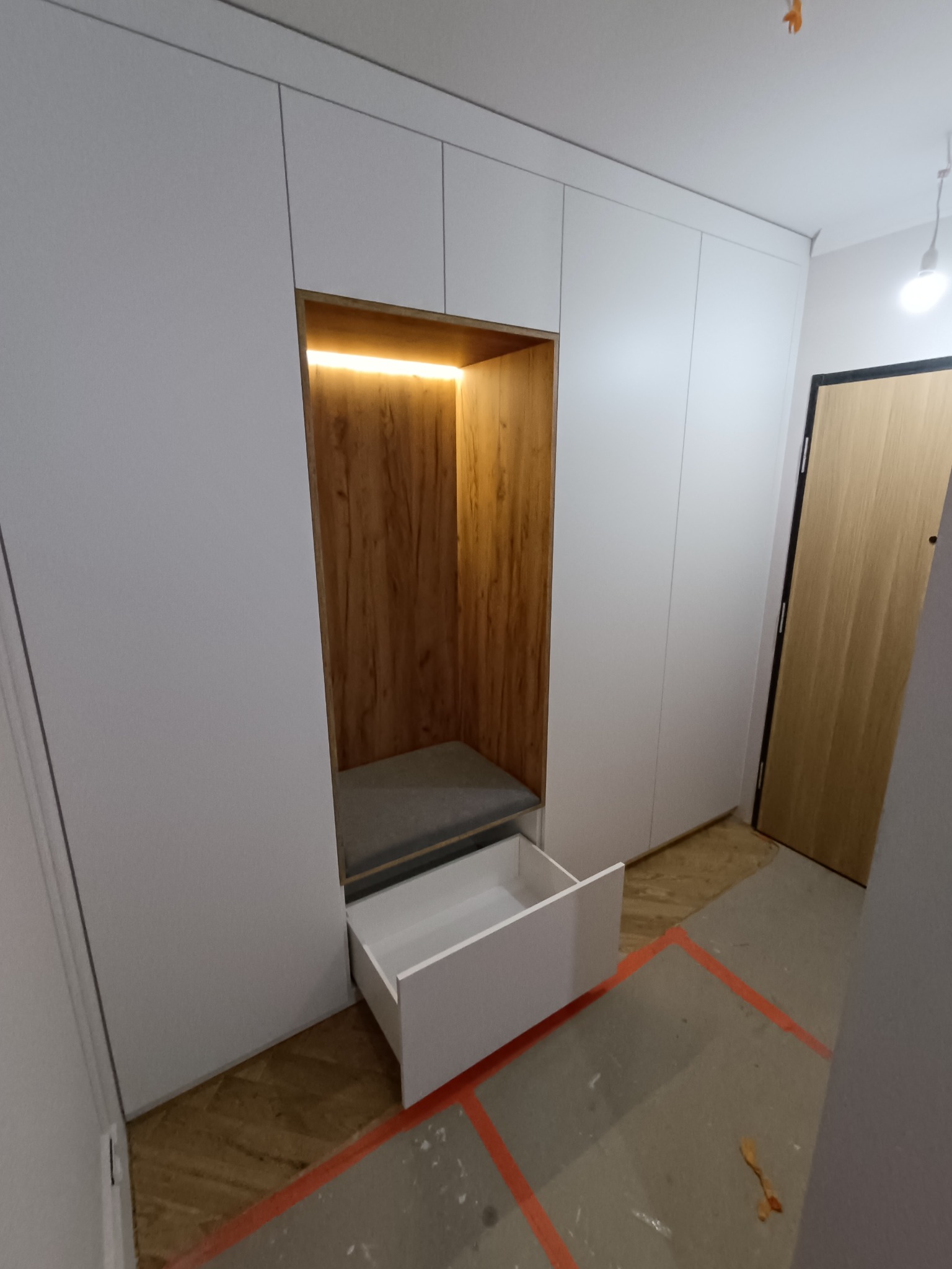 Biała szafa wnękowa z siedziskiem i wysuwaną szufladą, oświetlona ciepłym światłem. Widoczny fragment podłogi z parkietem i zabezpieczeniem w postaci taśmy.