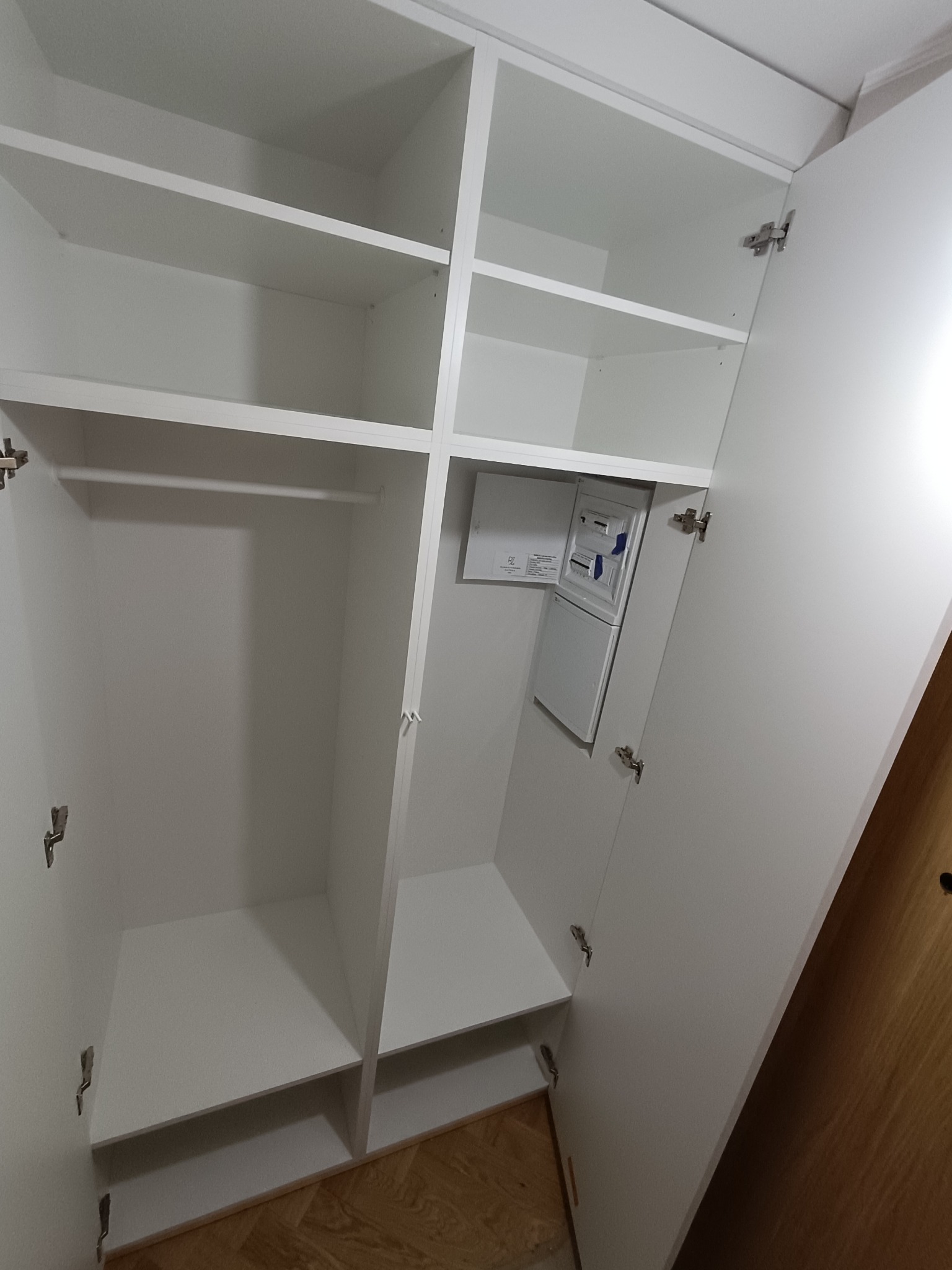 Biała szafa wnękowa z otwartymi drzwiami, widoczne półki i drążek na ubrania, zintegrowana skrzynka elektryczna. Minimalistyczny design, jasne wnętrze.