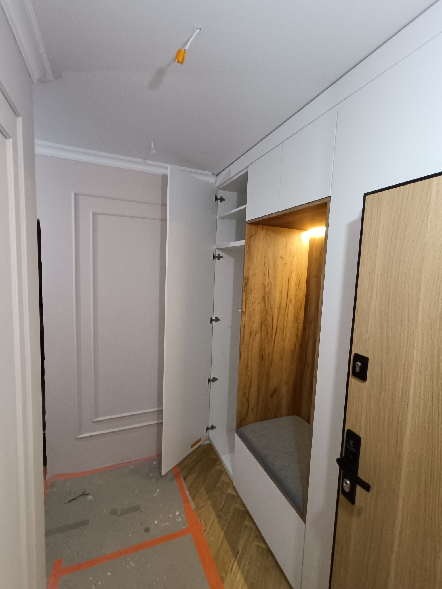 Biała szafa wnękowa z siedziskiem z drewnianym wykończeniem i oświetleniem, widok na otwarte drzwi szafy i fragment podłogi zabezpieczonej folią.