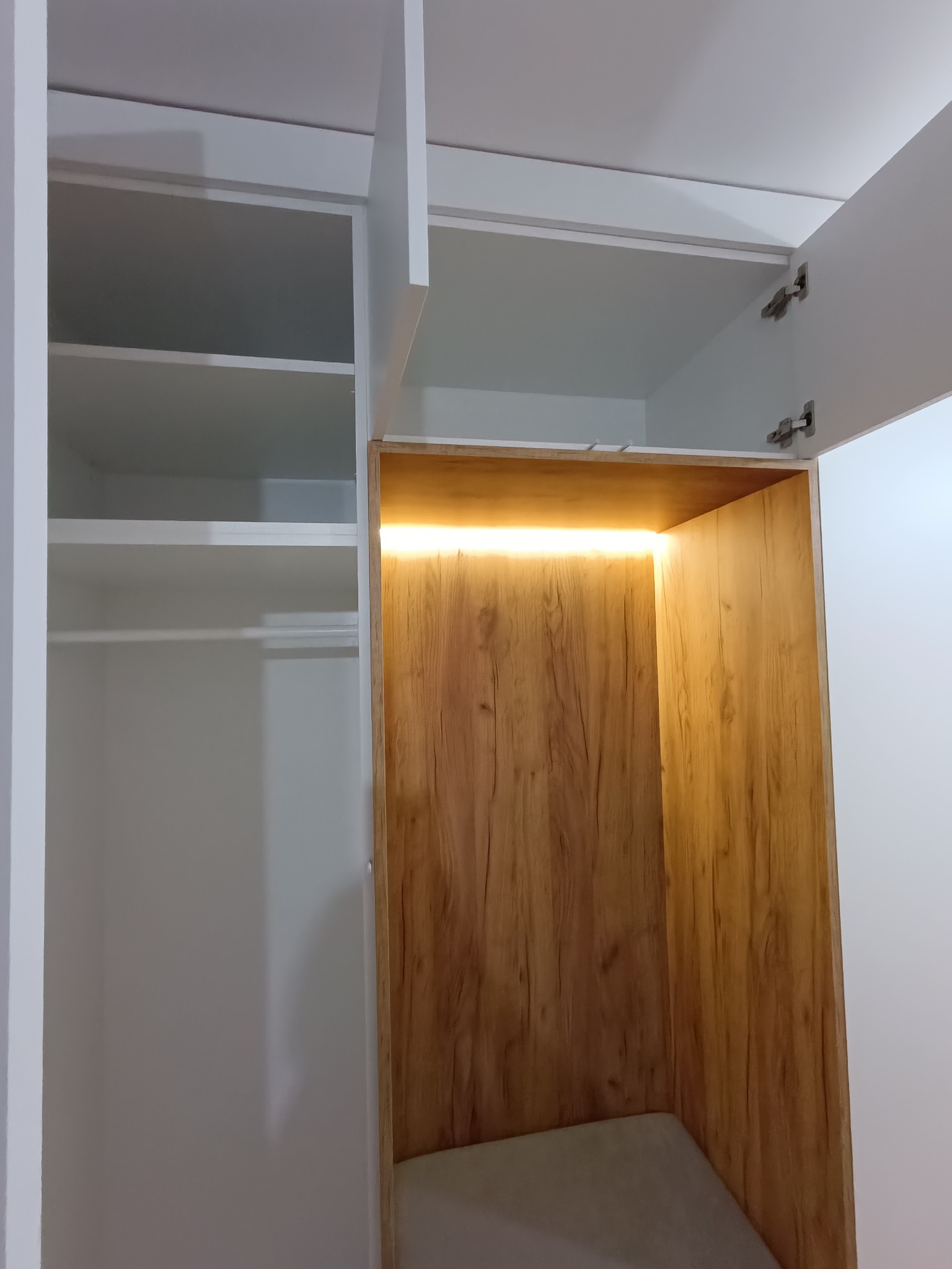 Wnętrze szafy z białymi półkami i drewnianą wnęką z oświetleniem LED. Drzwiczki szafy otwarte, widoczne zawiasy. Minimalistyczny design.
