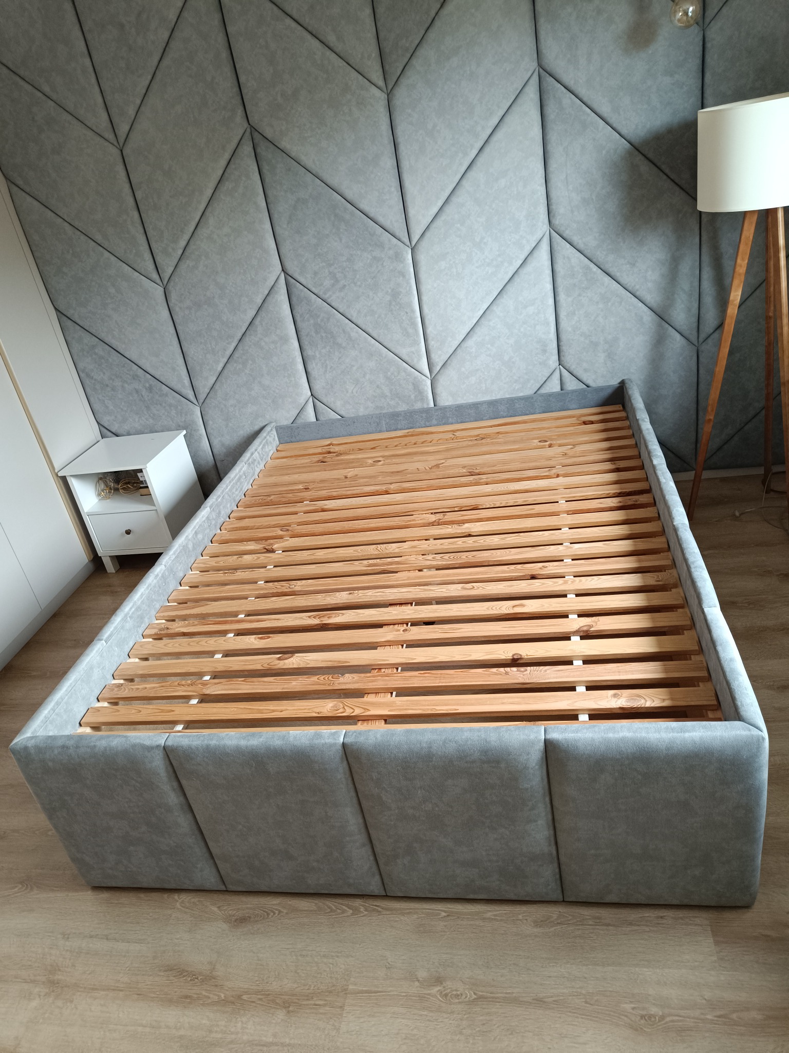 Szare łóżko tapicerowane bez materaca, z drewnianym stelażem, na tle ściany z geometrycznymi panelami. Obok szafka nocna i lampa podłogowa.