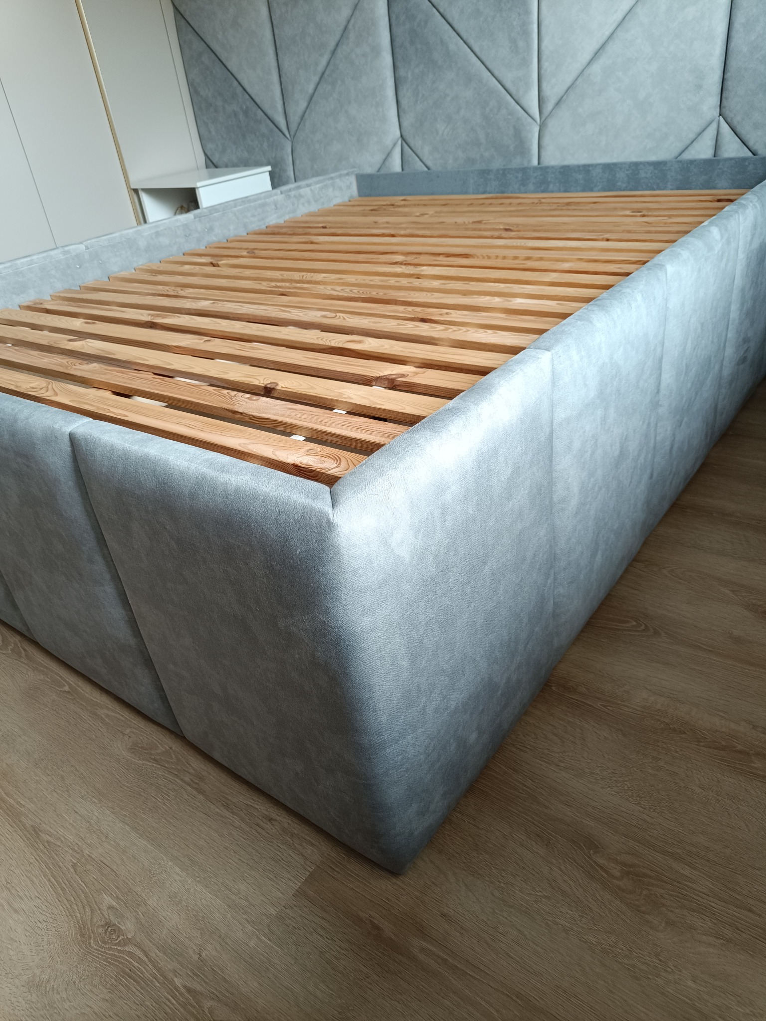 Szare, tapicerowane łóżko z drewnianym stelażem, widoczne z niskiego kąta. Wezgłowie z geometrycznym wzorem. Podłoga z paneli imitujących drewno.
