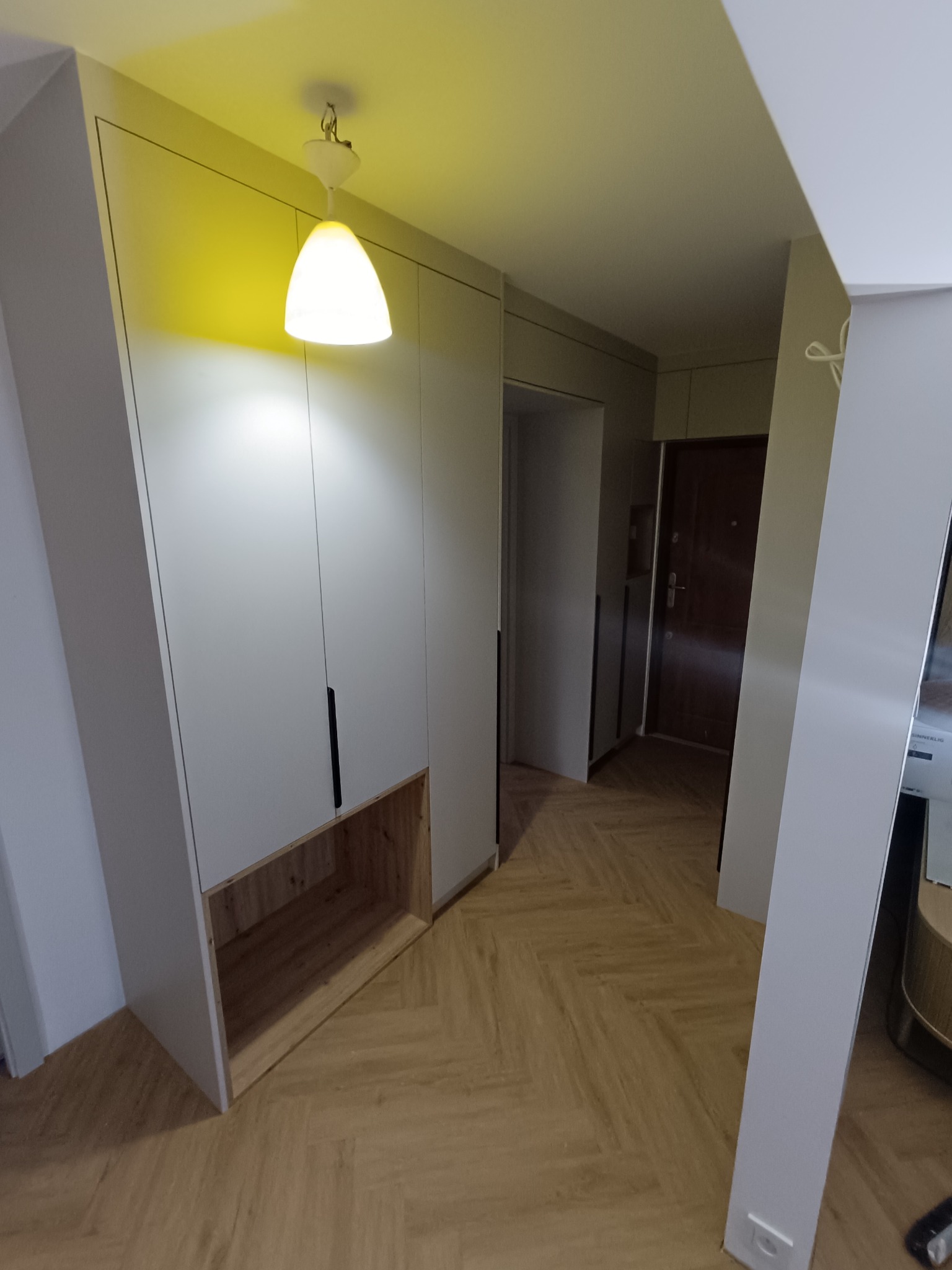 Biała szafa wnękowa z drewnianą półką w przedpokoju, oświetlona lampą sufitową. Parkiet w jodełkę na podłodze. Minimalistyczny design.