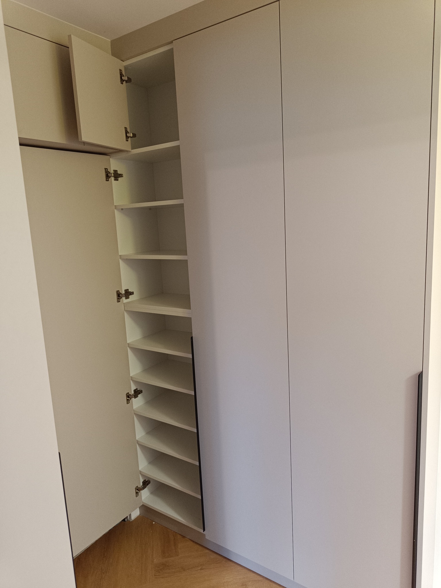 Jasnoszara szafa wnękowa z otwartymi drzwiami, ukazująca półki. Minimalistyczny design, widoczne zawiasy i drewniana podłoga w jodełkę. Ujęcie z bliska.