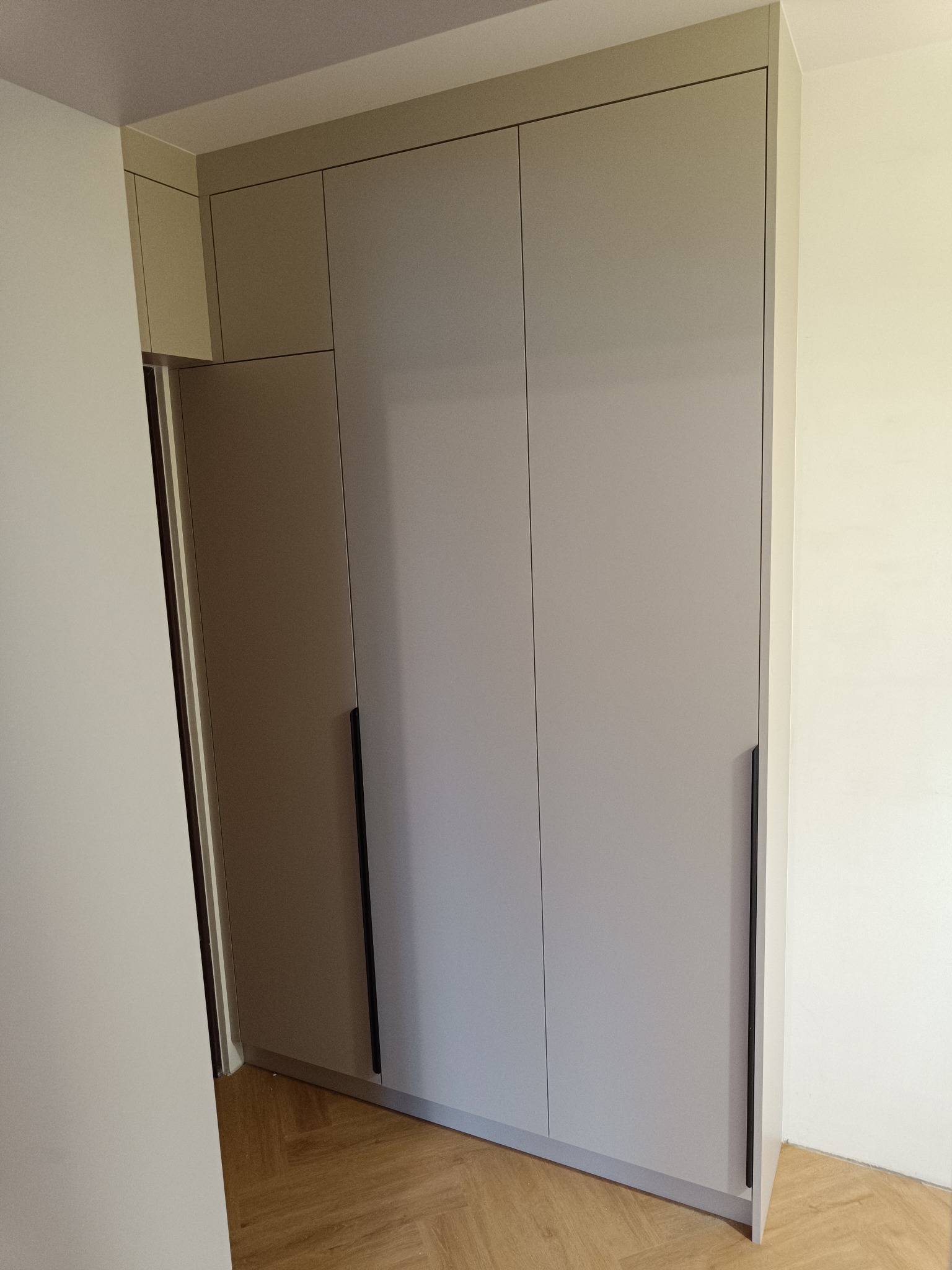 Jasnoszara szafa wnękowa z trzema drzwiami i długimi, czarnymi uchwytami, na parkiecie ułożonym w jodełkę. Widoczny fragment ściany i wejście do pomieszczenia.