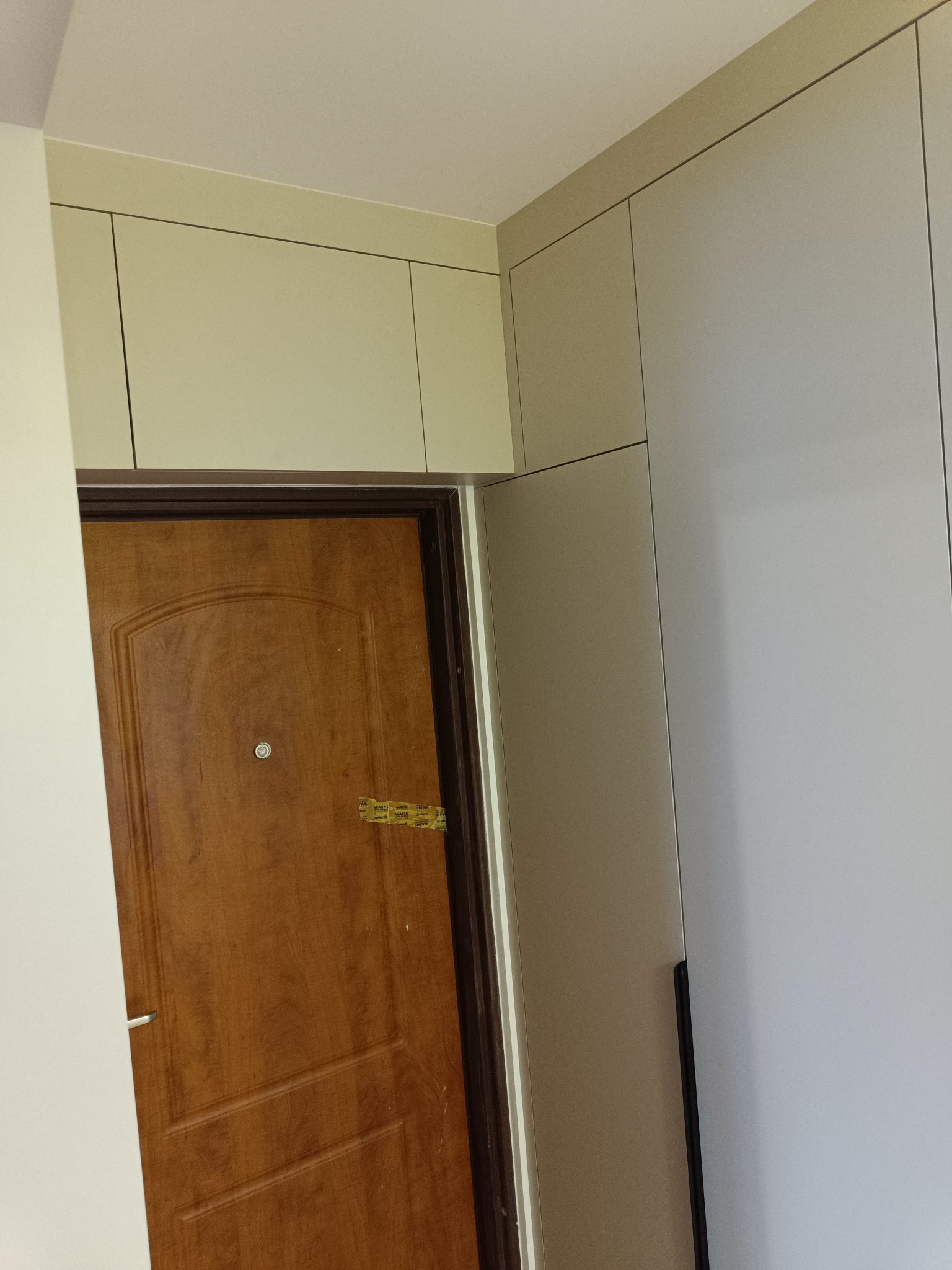 Jasna szafa wnękowa z frontami bez uchwytów, zabudowana nad brązowymi drzwiami wejściowymi. Minimalistyczny design, proste linie.