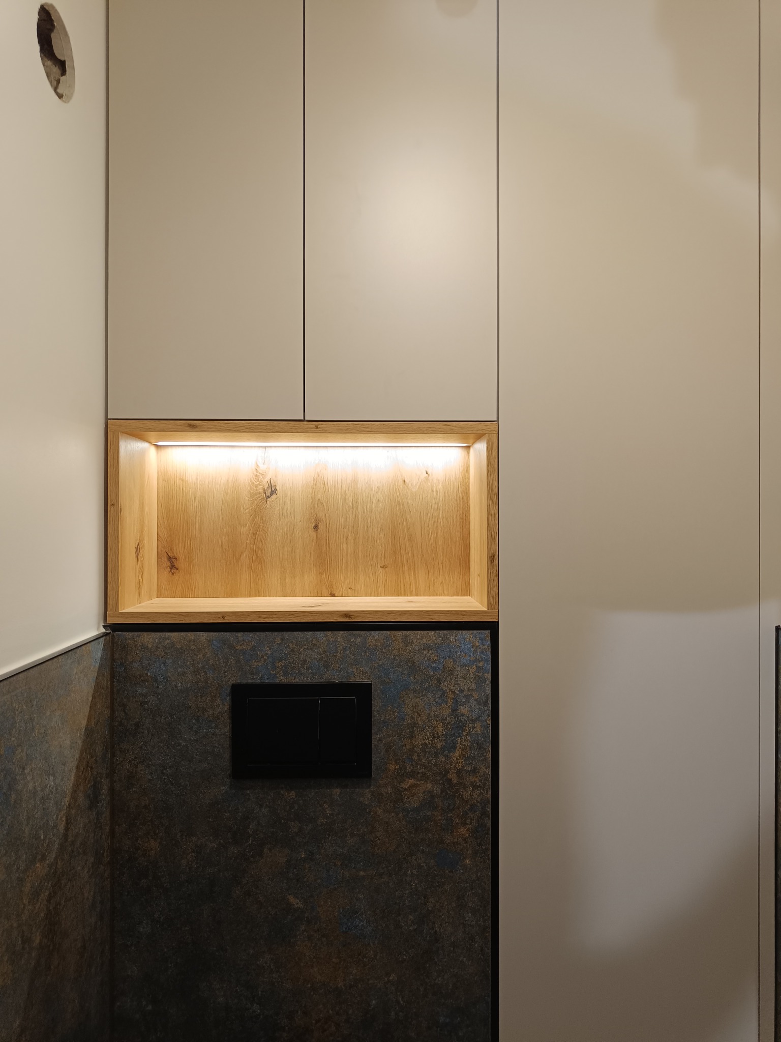 Beżowe szafki z drewnianą, podświetlaną wnęką nad panelem z przyciskiem spłuczki, na tle ściany z efektem rdzy. Minimalistyczny design.