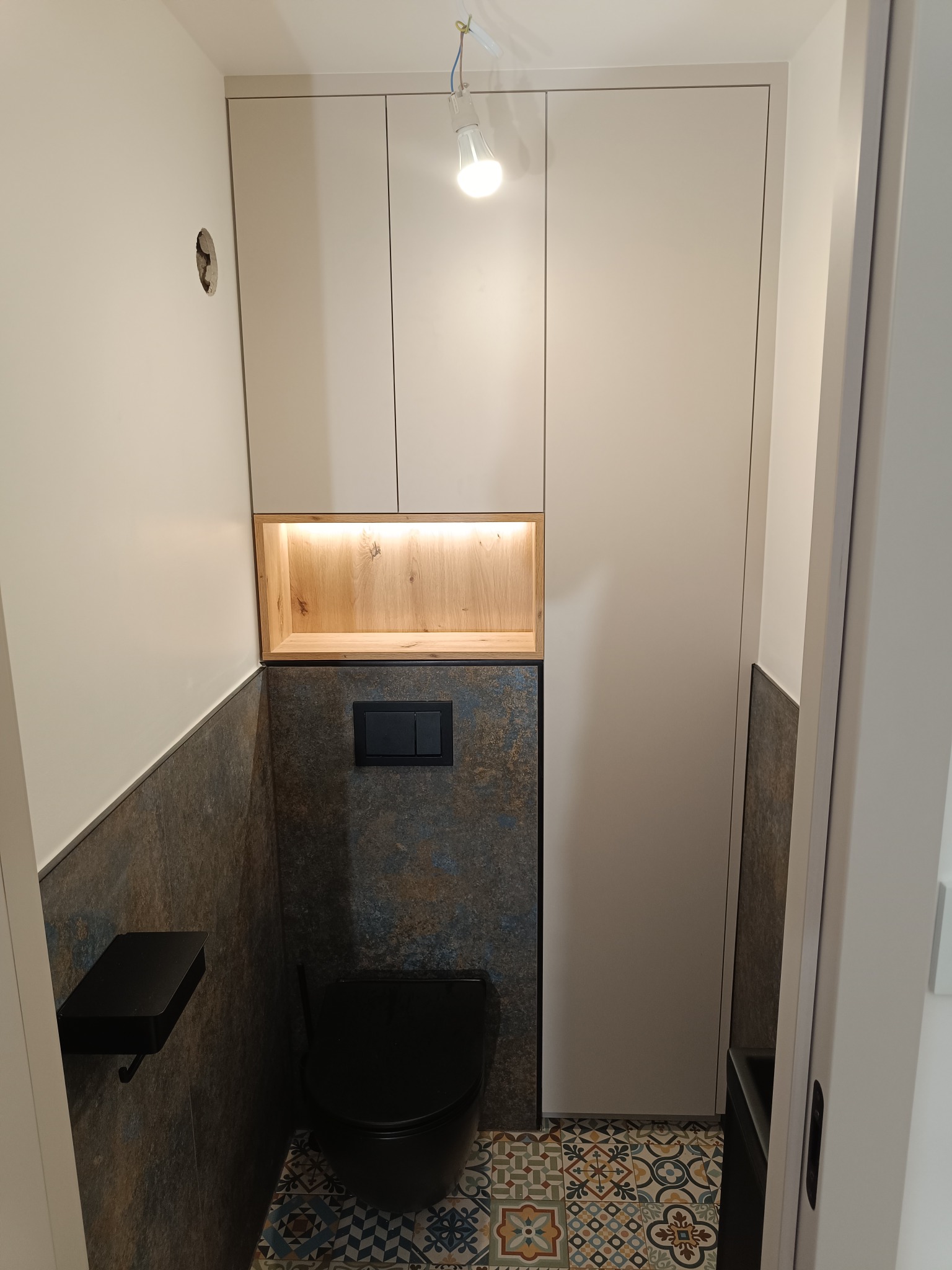 Wąska toaleta z szafkami na wymiar w zabudowie, podświetlana półka z drewnianym wykończeniem, czarna armatura i wzorzysta podłoga. Minimalistyczny design.