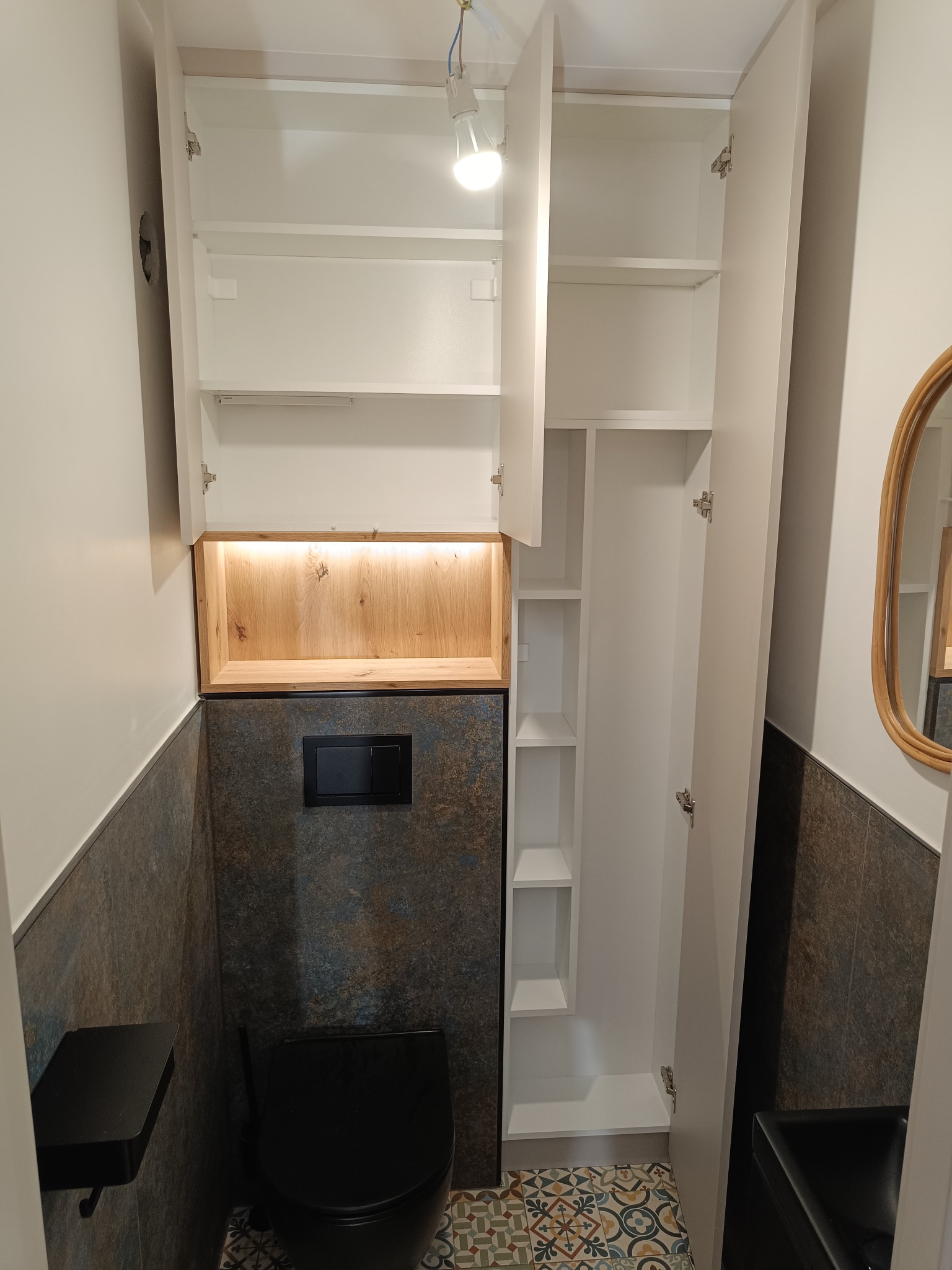 Biała szafa wnękowa z oświetleniem LED w drewnianej obudowie, otwarte drzwi ukazują półki. U dołu widoczna toaleta i wzorzysta podłoga.