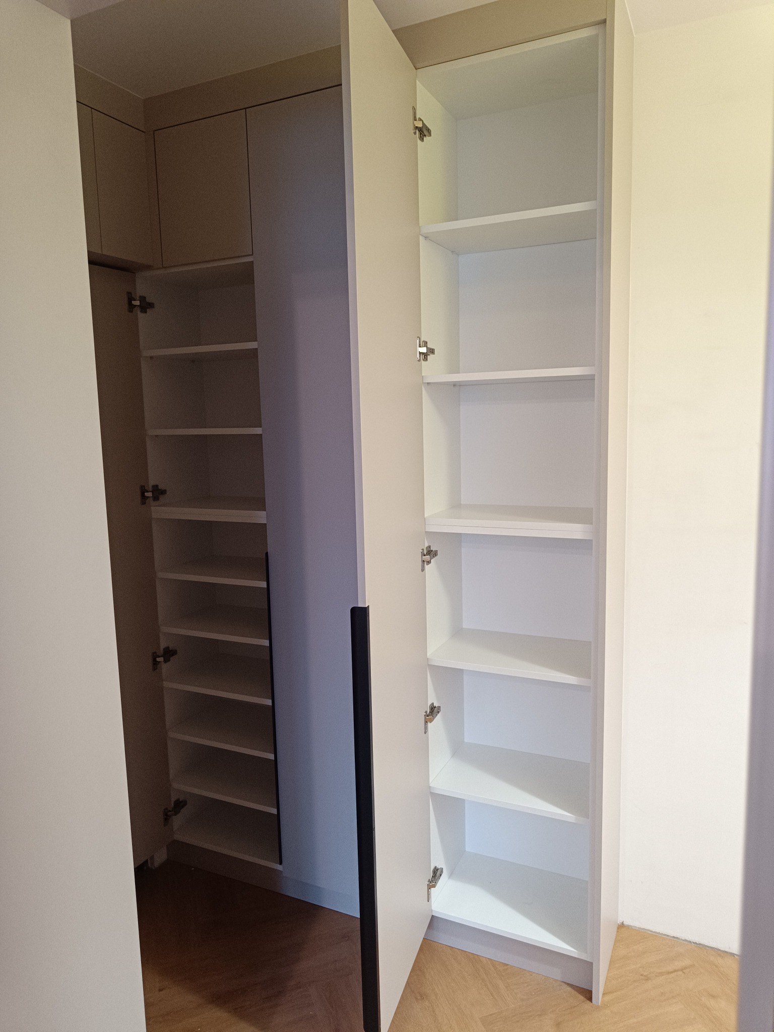 Otwarta, biała szafa wnękowa z półkami, widoczna z perspektywy wnętrza. Minimalistyczny design, jasne oświetlenie, drewniana podłoga. Drzwi szafy z czarnym, pionowym uchwytem.